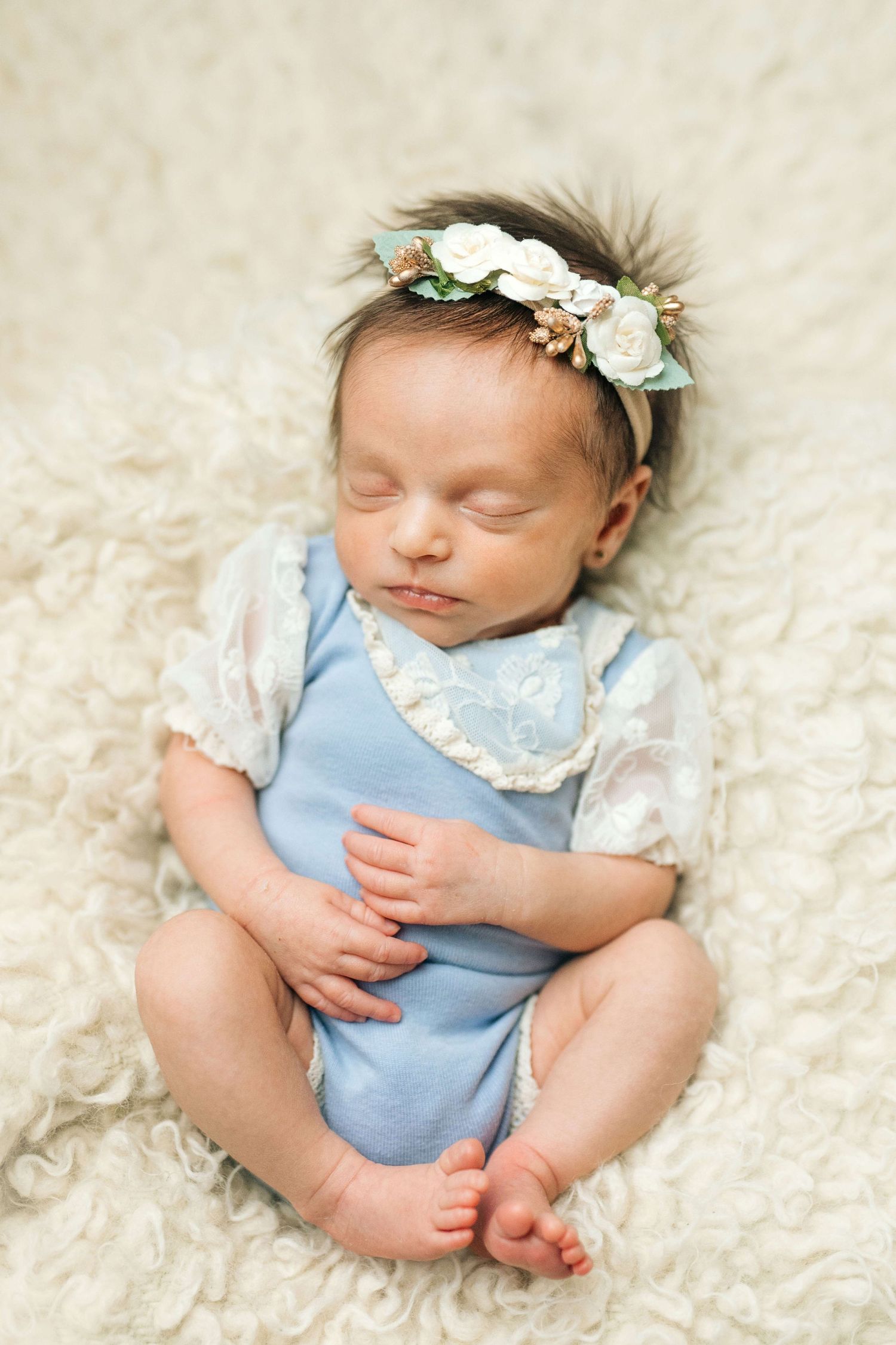 Sleeping newborn baby girl in blue romper and floral headband on cream fluffy blanket.