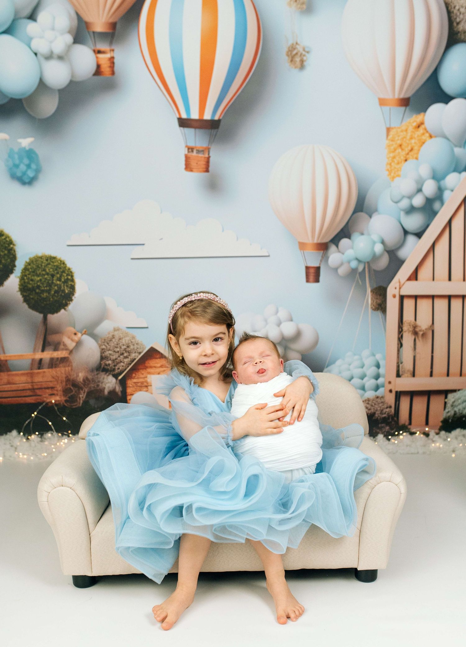A young girl in a blue tulle dress holds a newborn on a small sofa with hot air balloon backdrop.