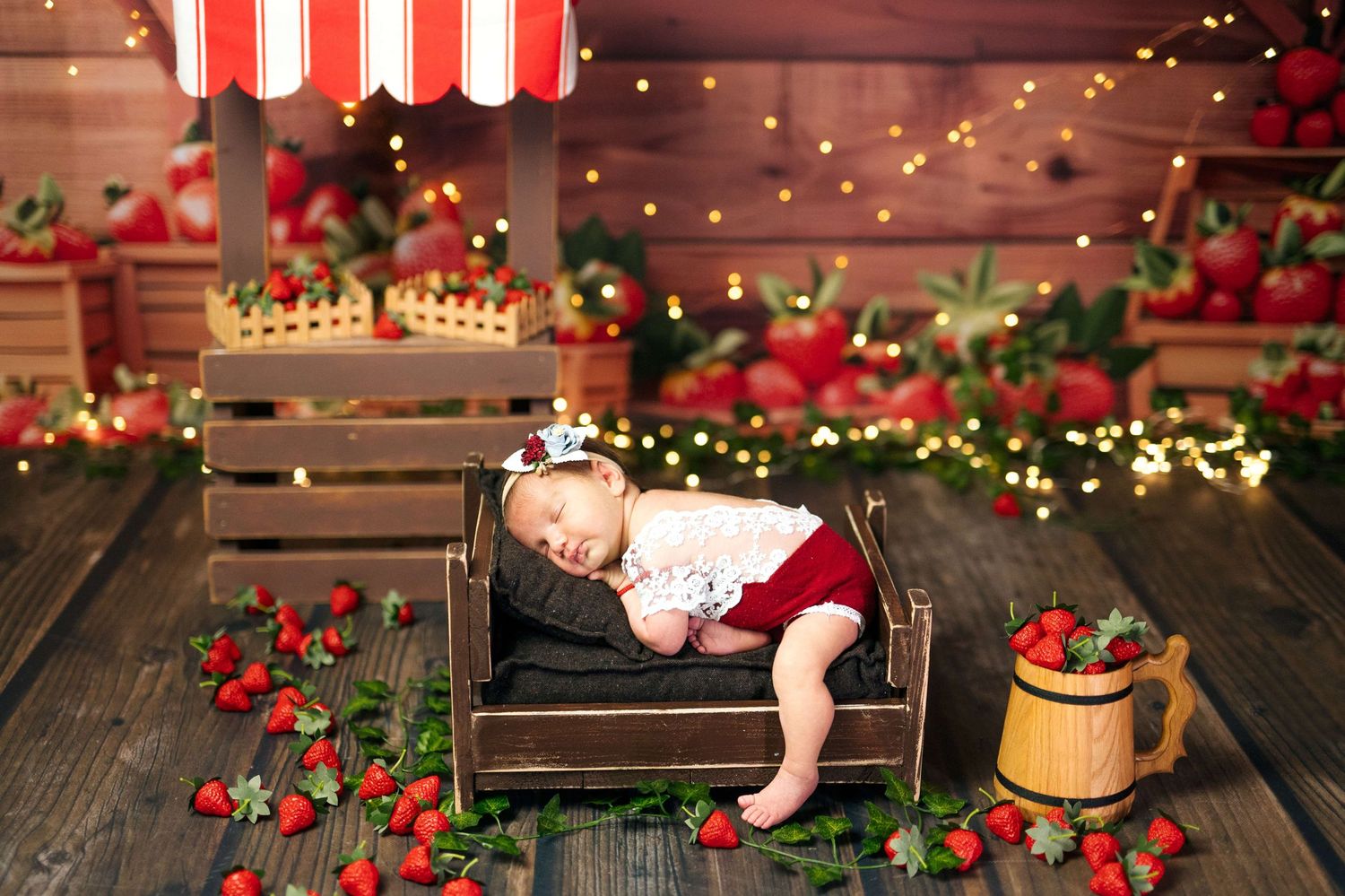 Newborn baby sleeping in a tiny wooden bed surrounded by fresh strawberries and fairy lights in a charming market backdrop.