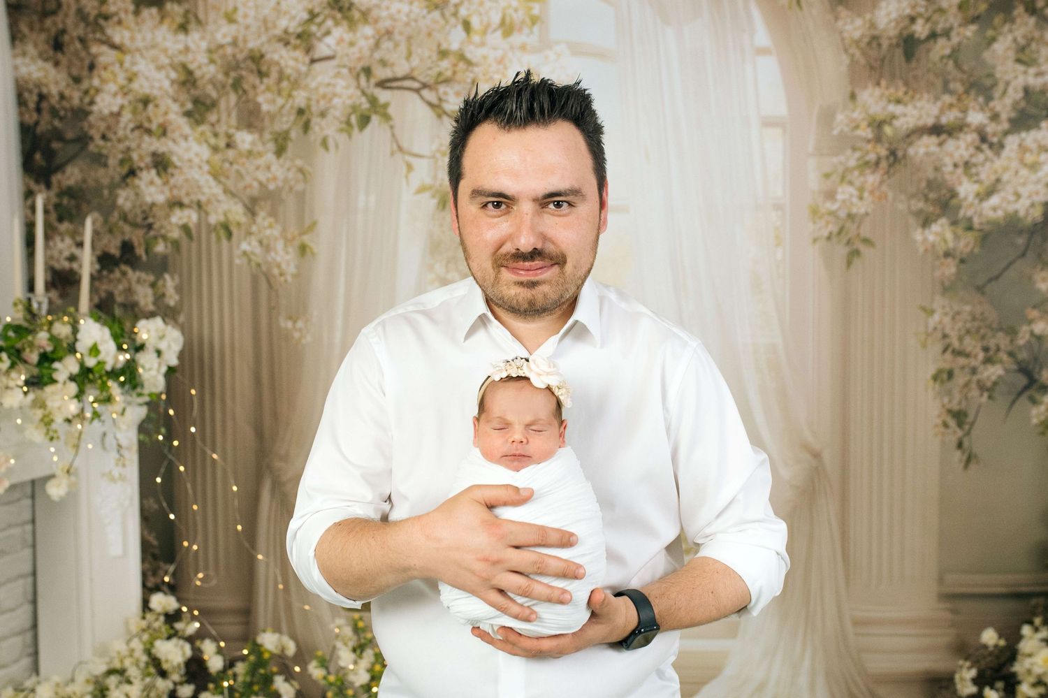 A father in a white shirt gently cradles his sleeping newborn baby amid beautiful white floral decor.