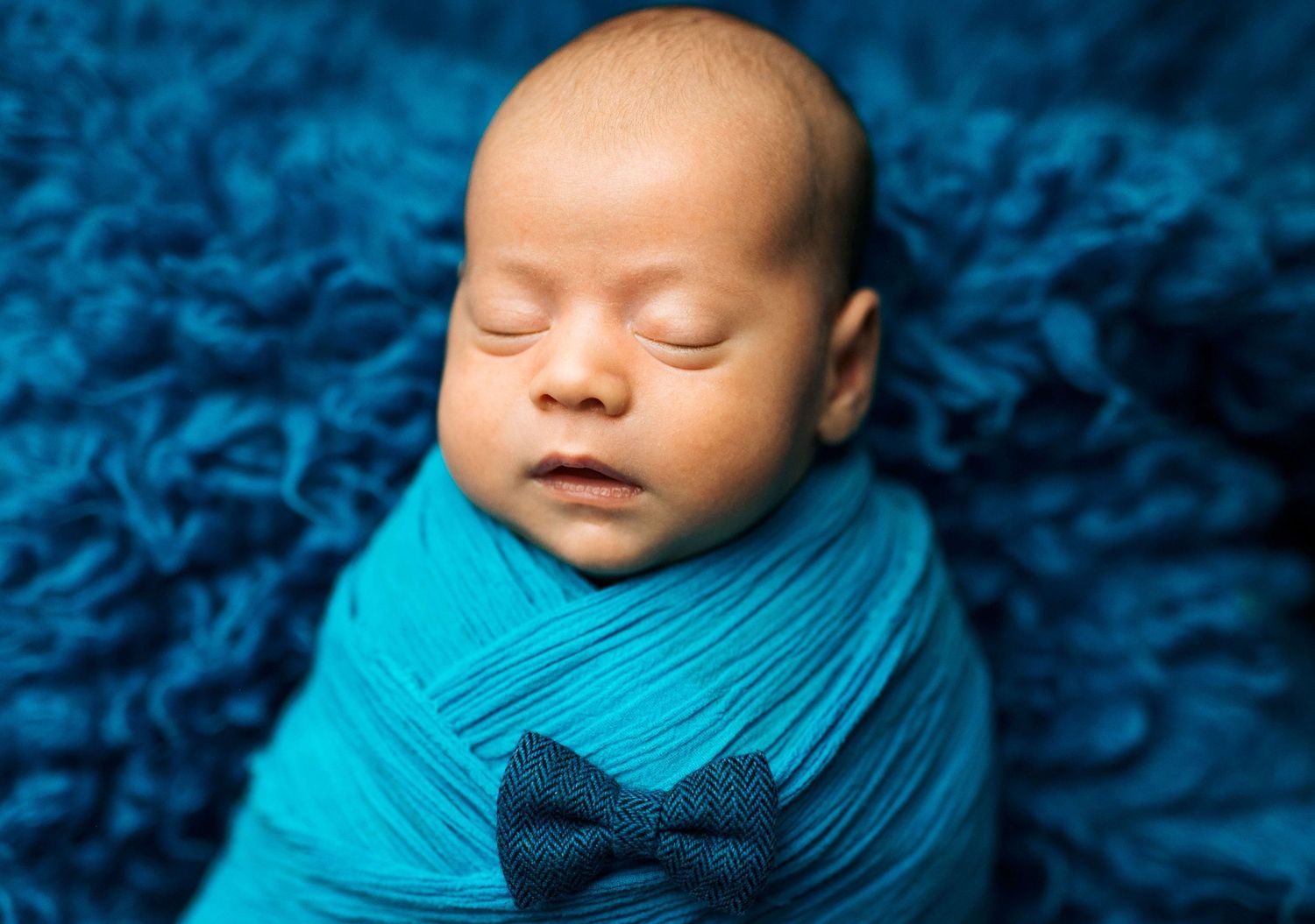 Sleeping newborn baby swaddled in teal wrap with navy bow tie on blue fluffy blanket.