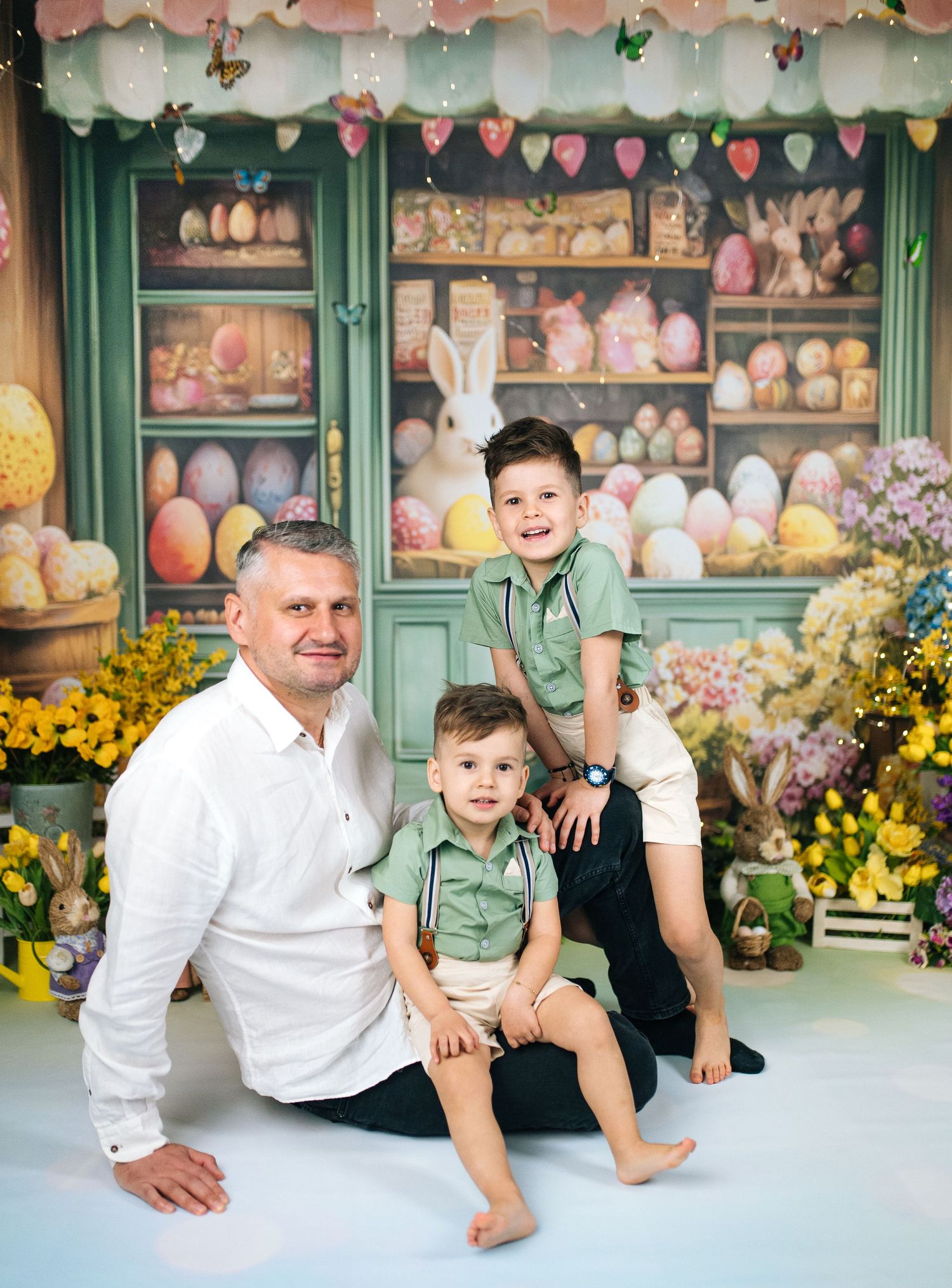 A man and two young boys pose together in front of a colorful Easter-themed backdrop with eggs and bunnies.