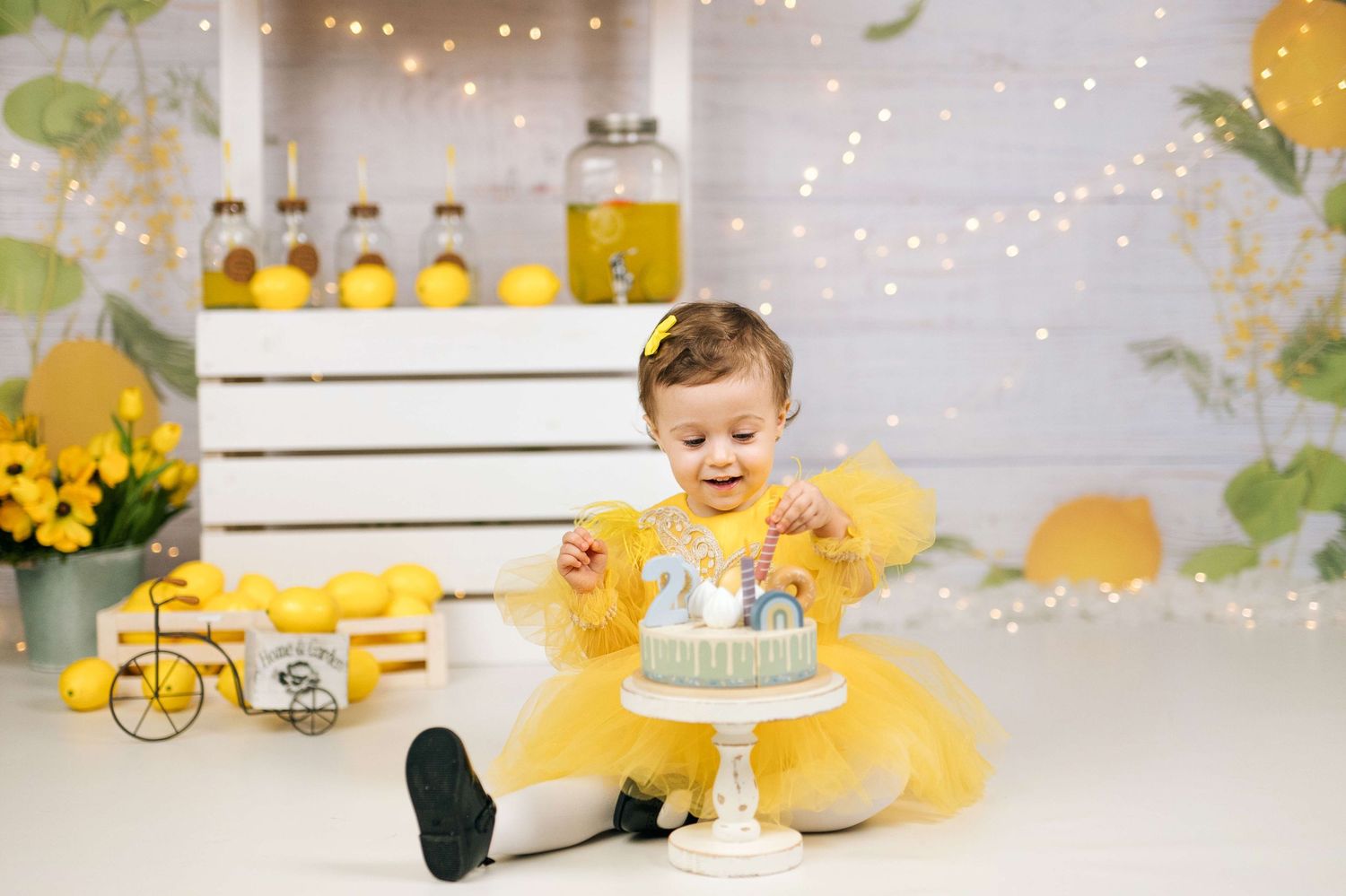 Baby girl in yellow tutu dress reaches for gray first birthday cake surrounded by lemons and fairy lights.