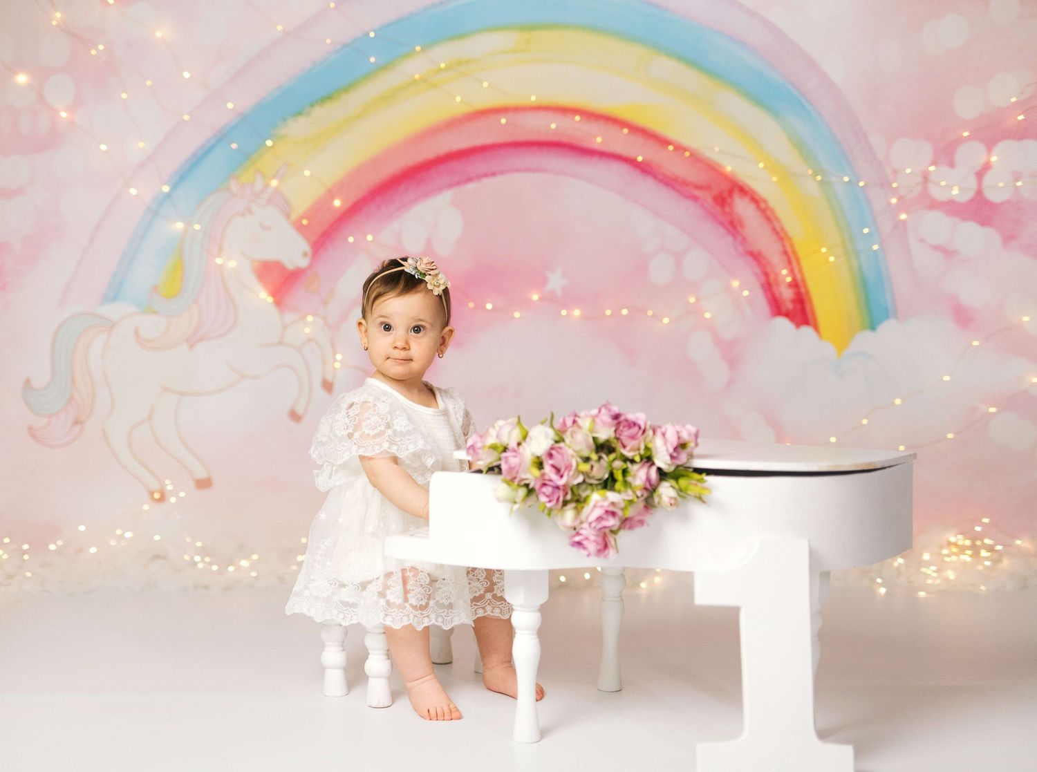 Toddler girl in white lace dress poses by mini white piano with flowers, rainbow unicorn backdrop.