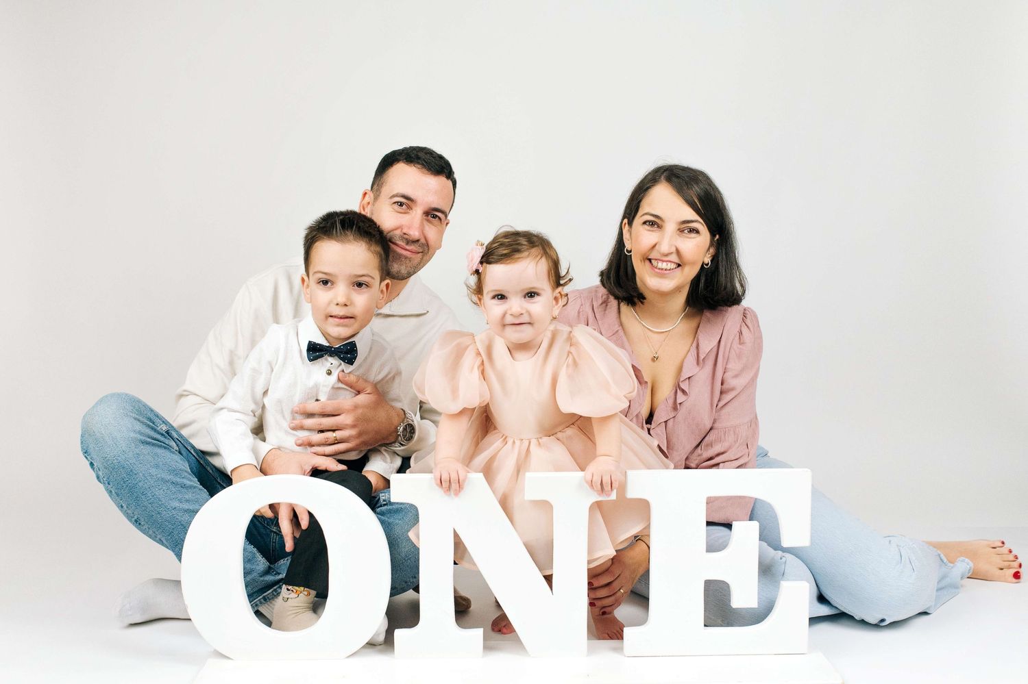 Family of four posing with ONE letter blocks celebrating baby girl's first birthday in white studio.