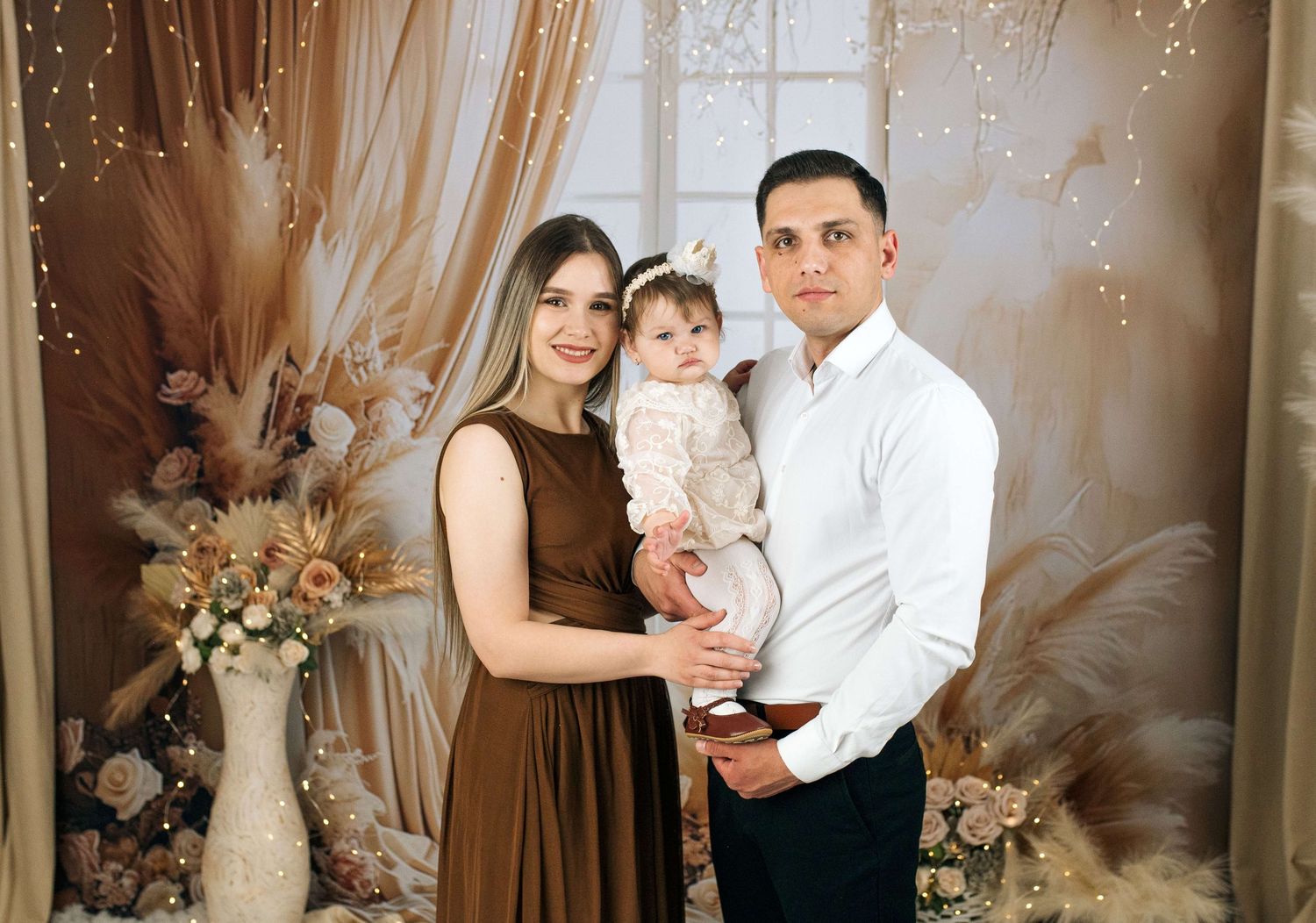 A young family poses together in a boho-styled photo shoot, with a mother, father, and baby girl in elegant attire.