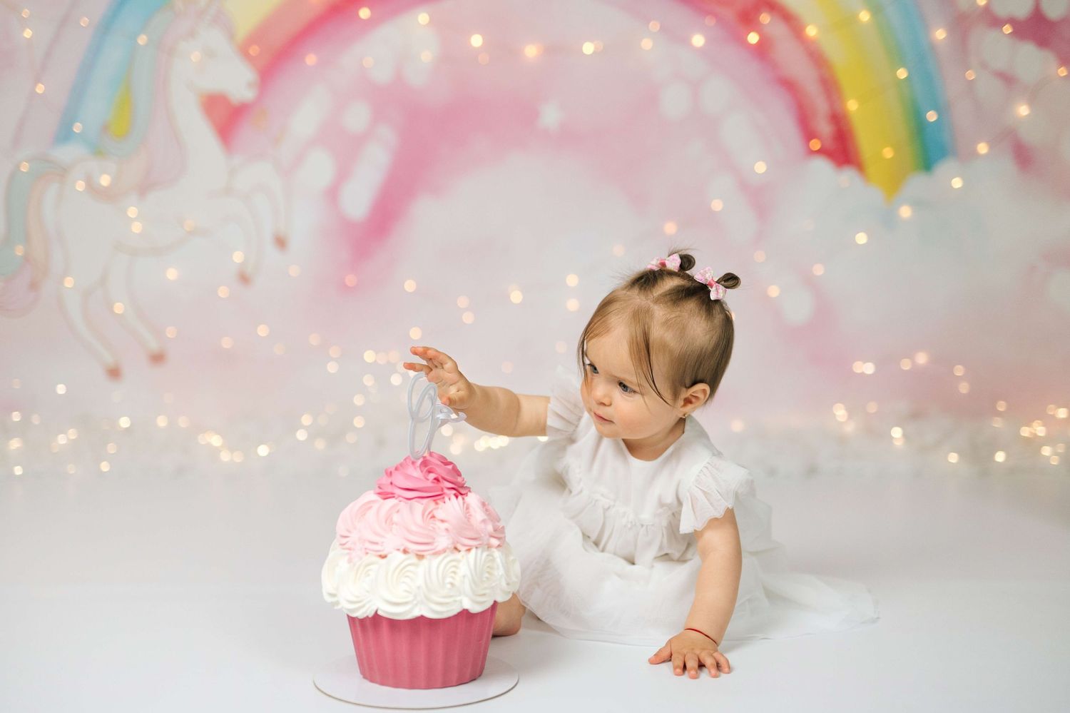 Baby girl in white dress touches pink giant cupcake during first birthday cake smash with rainbow backdrop.