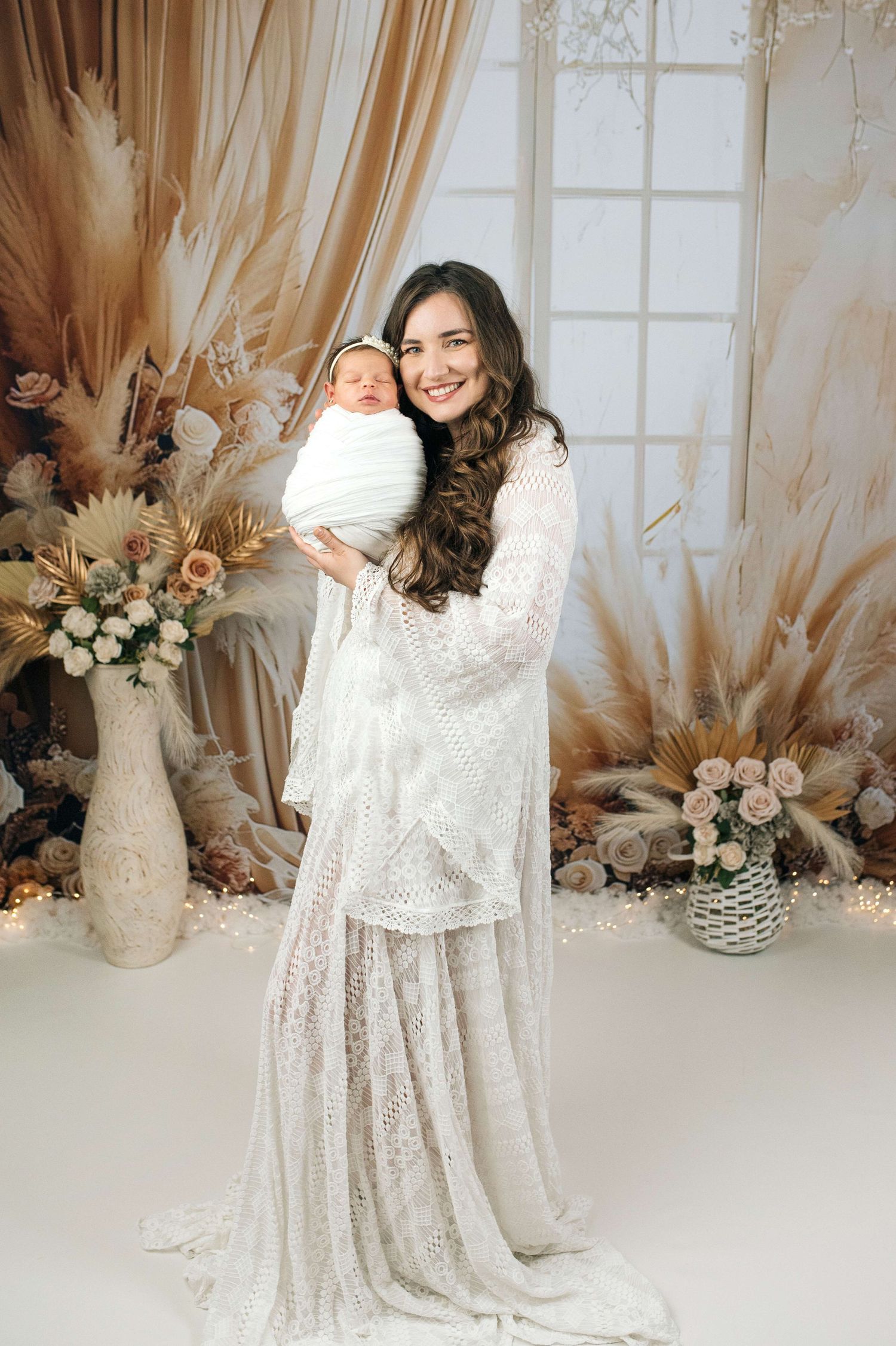 A smiling woman in a white lace gown holds her swaddled newborn in a boho-styled studio with floral decor.