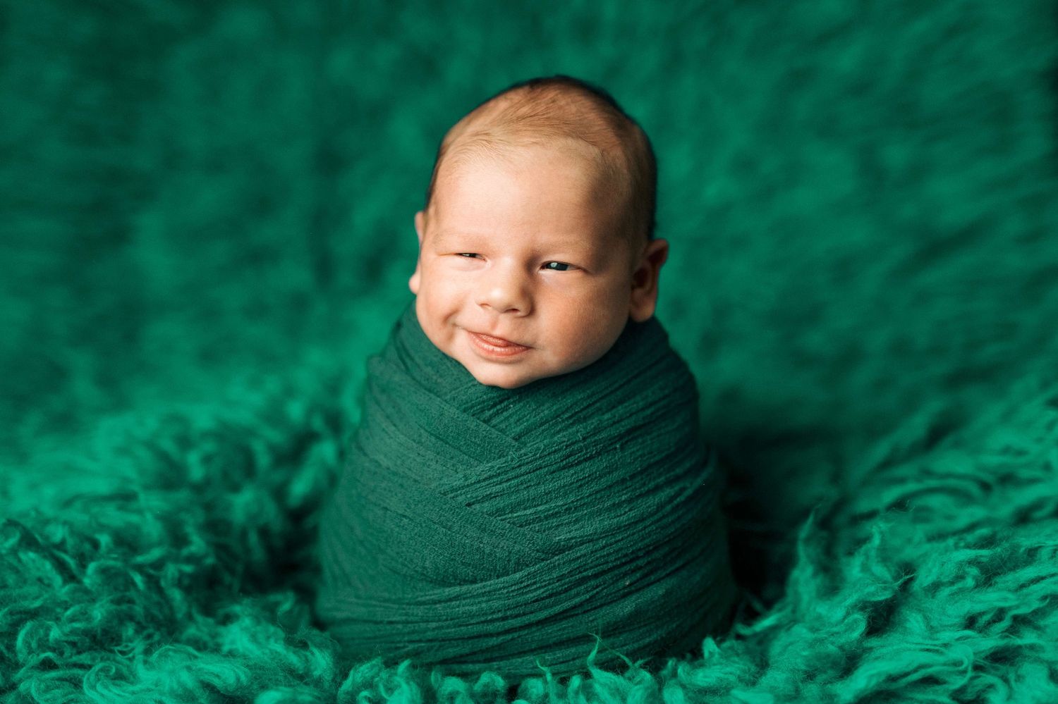 Smiling newborn baby wrapped in green swaddle on fluffy emerald green blanket for professional newborn photography.