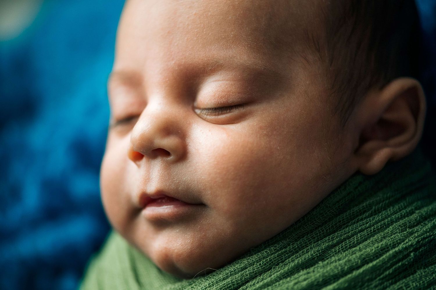 Peaceful newborn baby sleeping soundly, swaddled in a green wrap against a blue background.