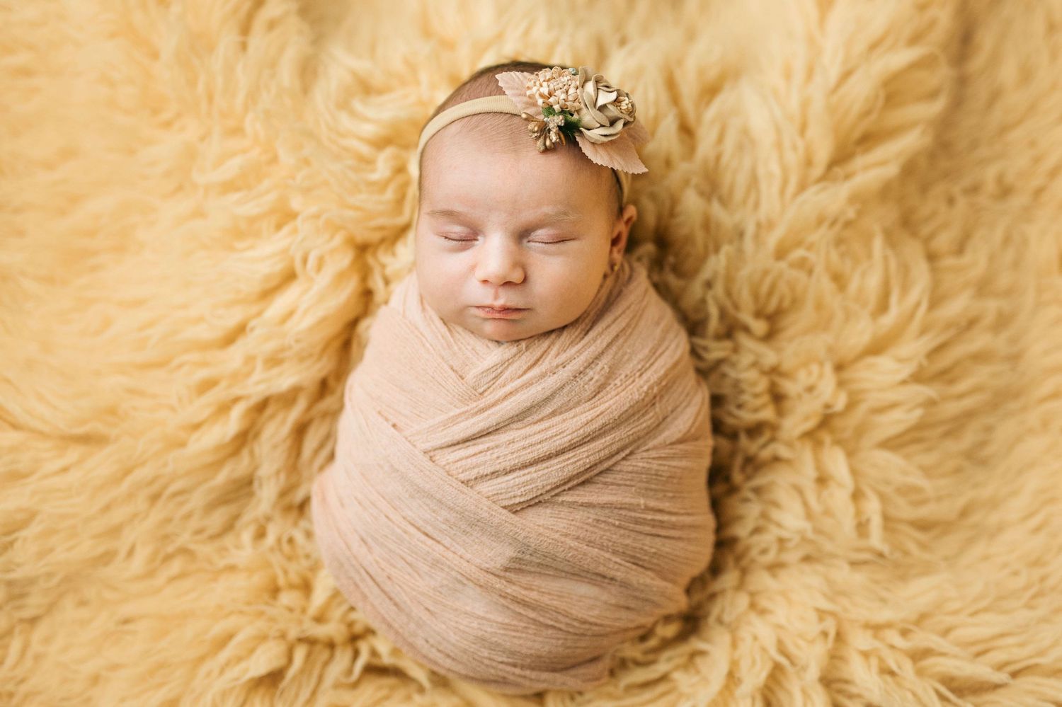 Newborn baby swaddled in peach wrap with floral headband, sleeping peacefully on soft yellow fluffy blanket.