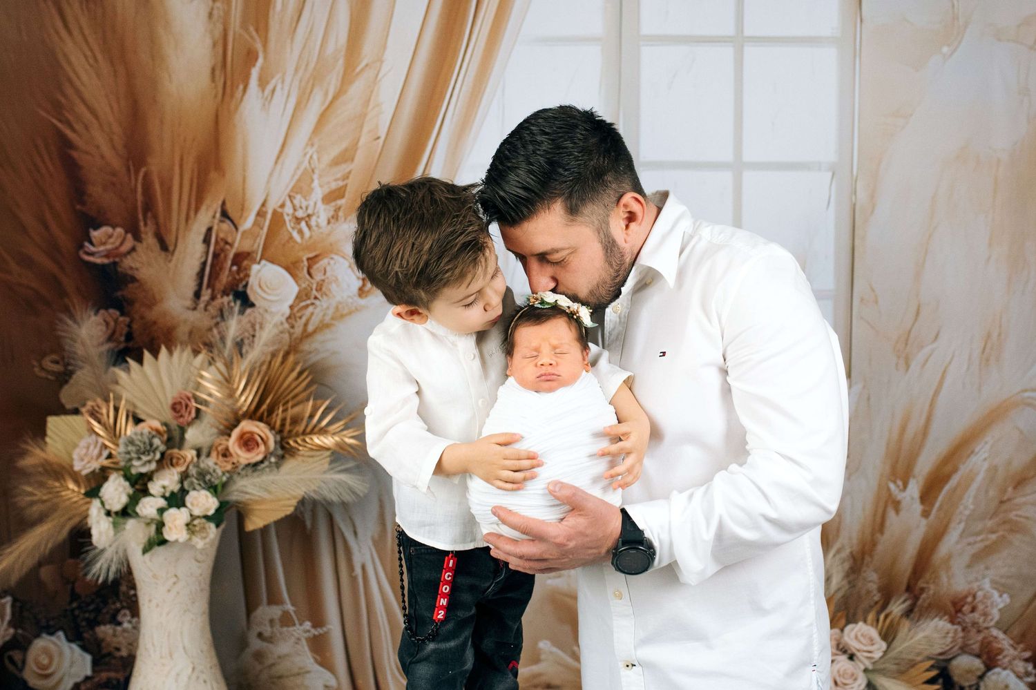 Father holding newborn baby with young son beside him in a warm, elegant studio portrait setting.