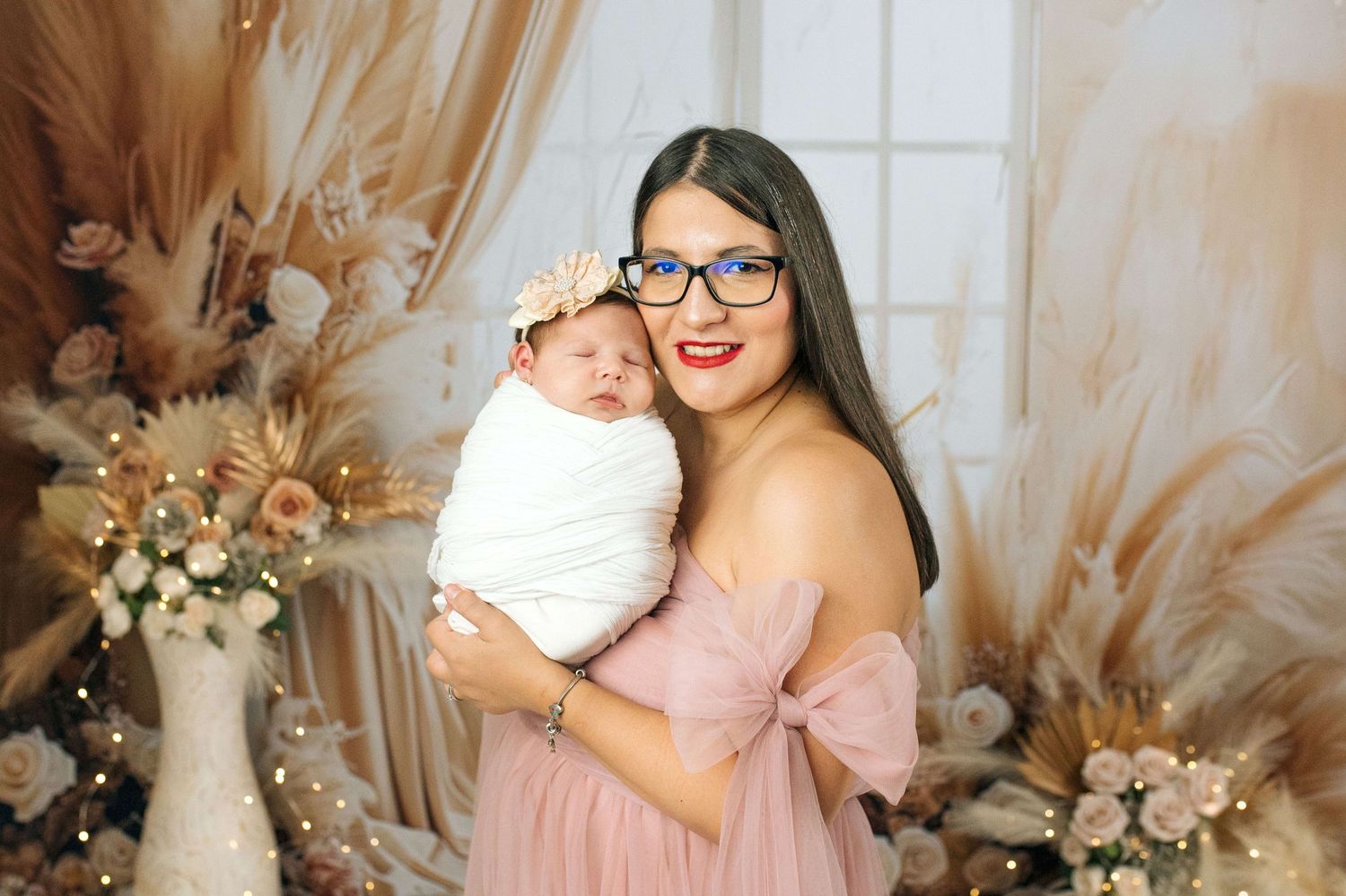 A young mother in a pink dress and glasses tenderly holds her newborn baby wrapped in white.