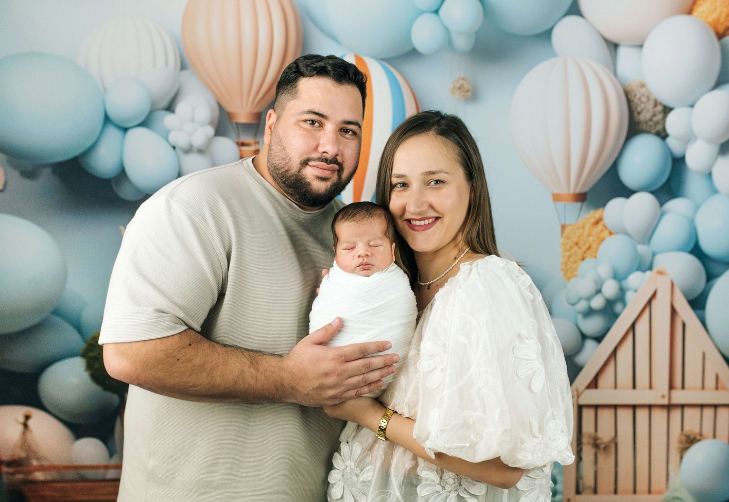 A couple smiles holding their newborn baby in front of a blue balloon and hot air balloon themed backdrop.