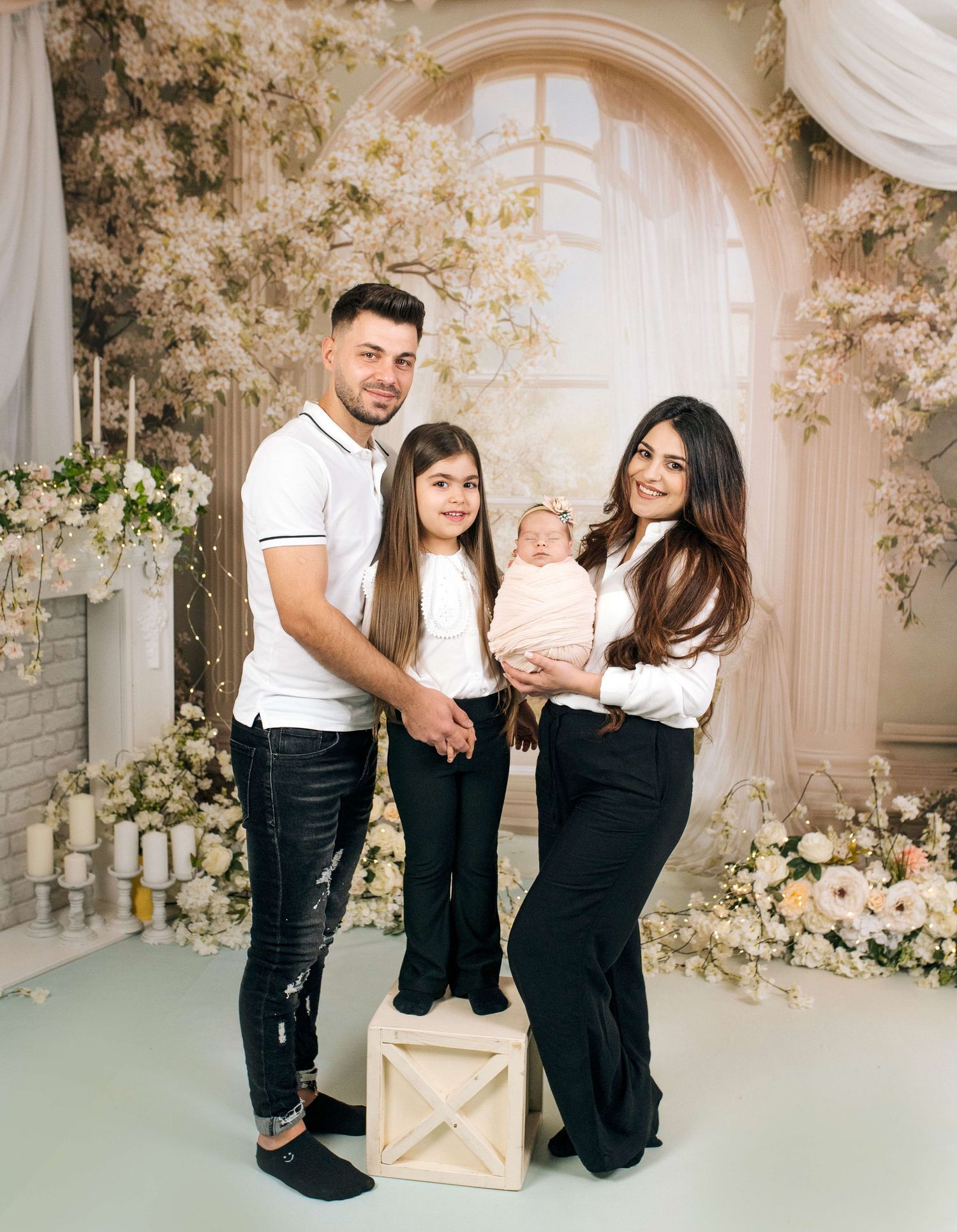 Family of four posing in floral studio, parents in white tops and black pants with young girl and newborn baby.