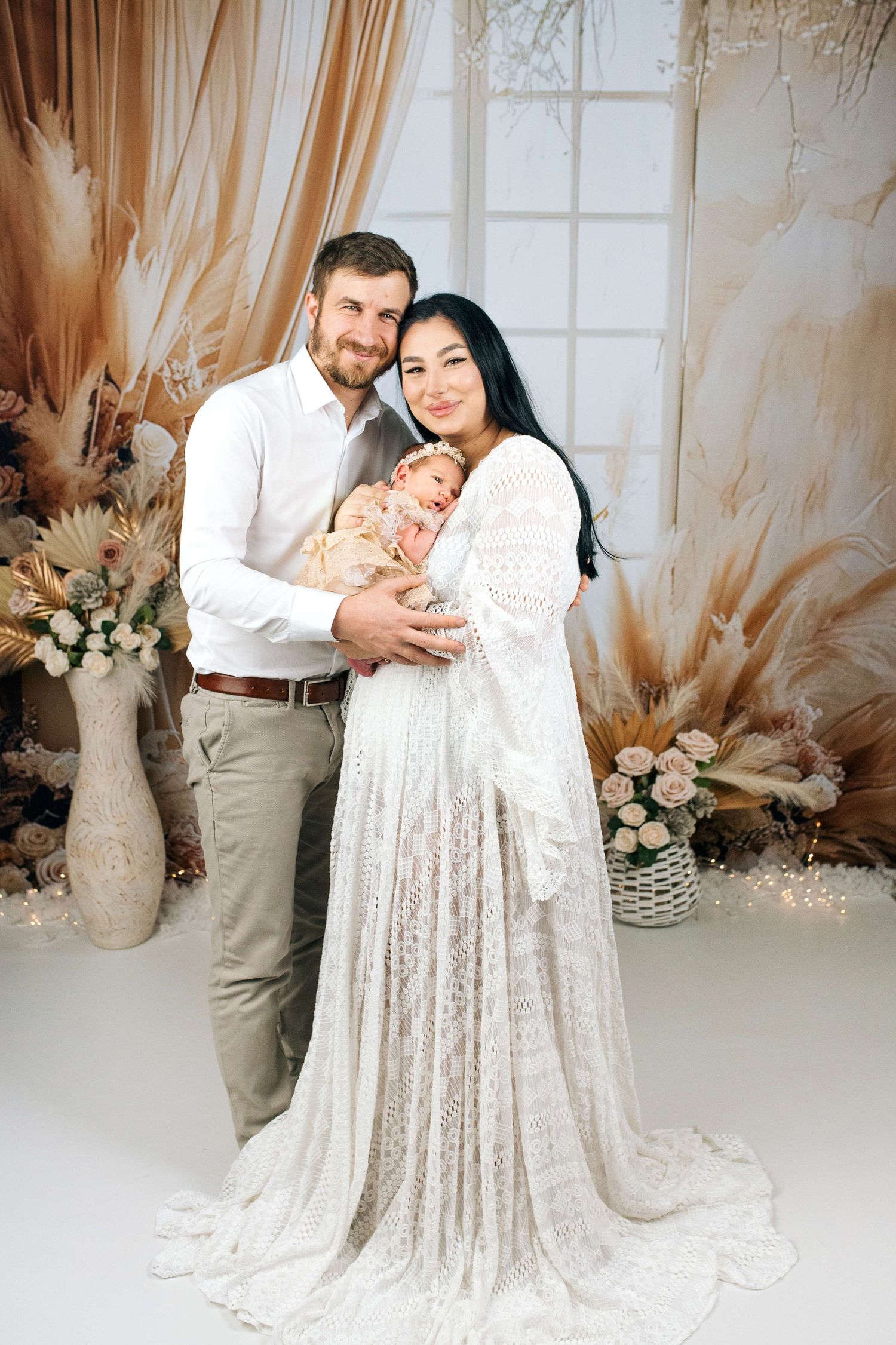 Happy couple in white and beige attire holding newborn baby in floral boho studio portrait.