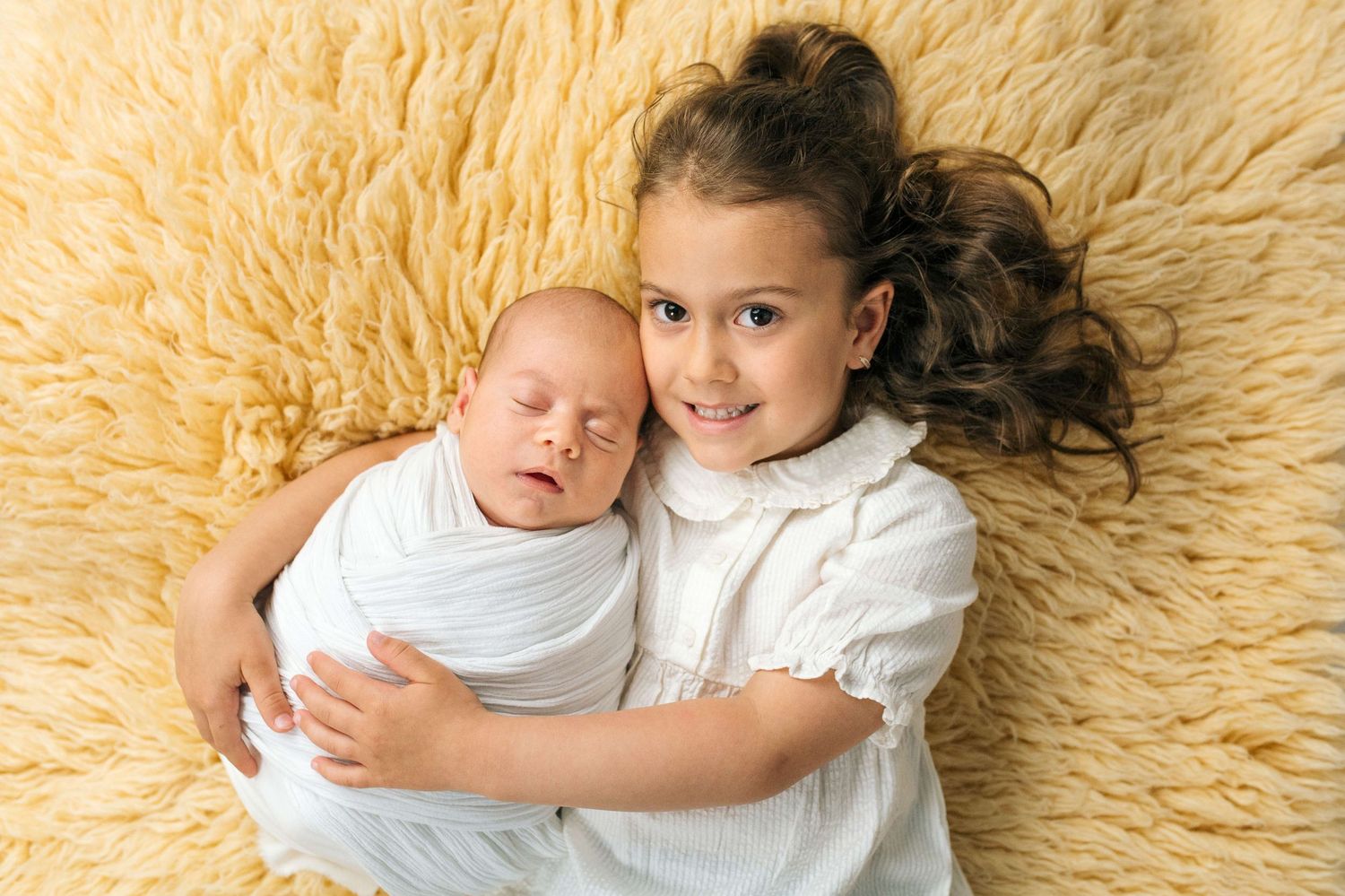 A young girl smiles while gently holding a swaddled newborn baby on a soft cream fluffy rug.