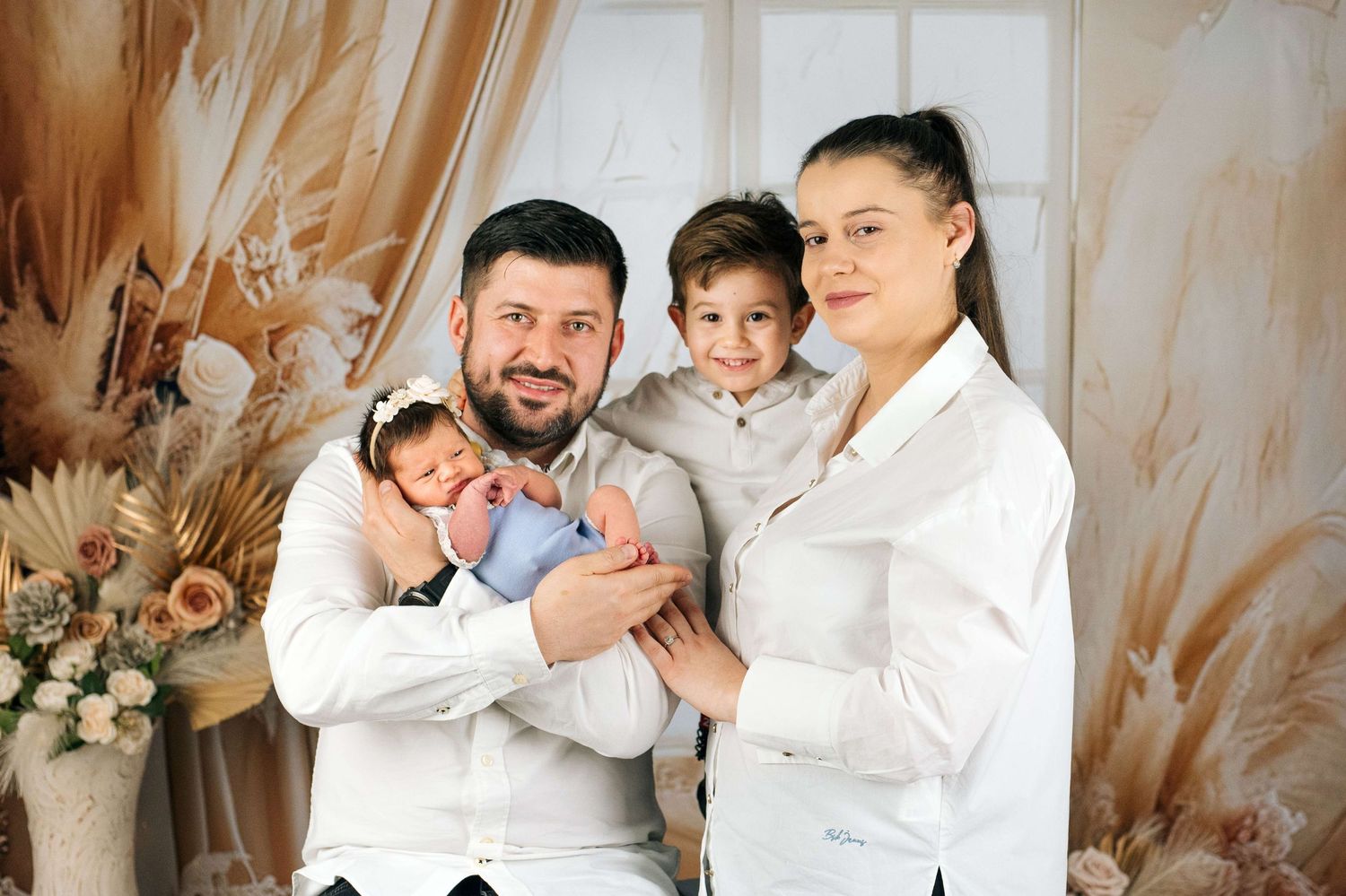 Family portrait with parents in white shirts holding newborn baby and young son in a decorated studio.