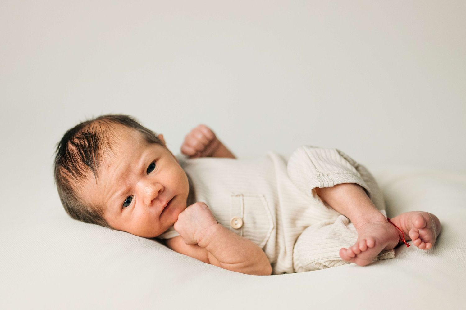Awake newborn baby in cream romper lying on white backdrop during professional newborn photography session.