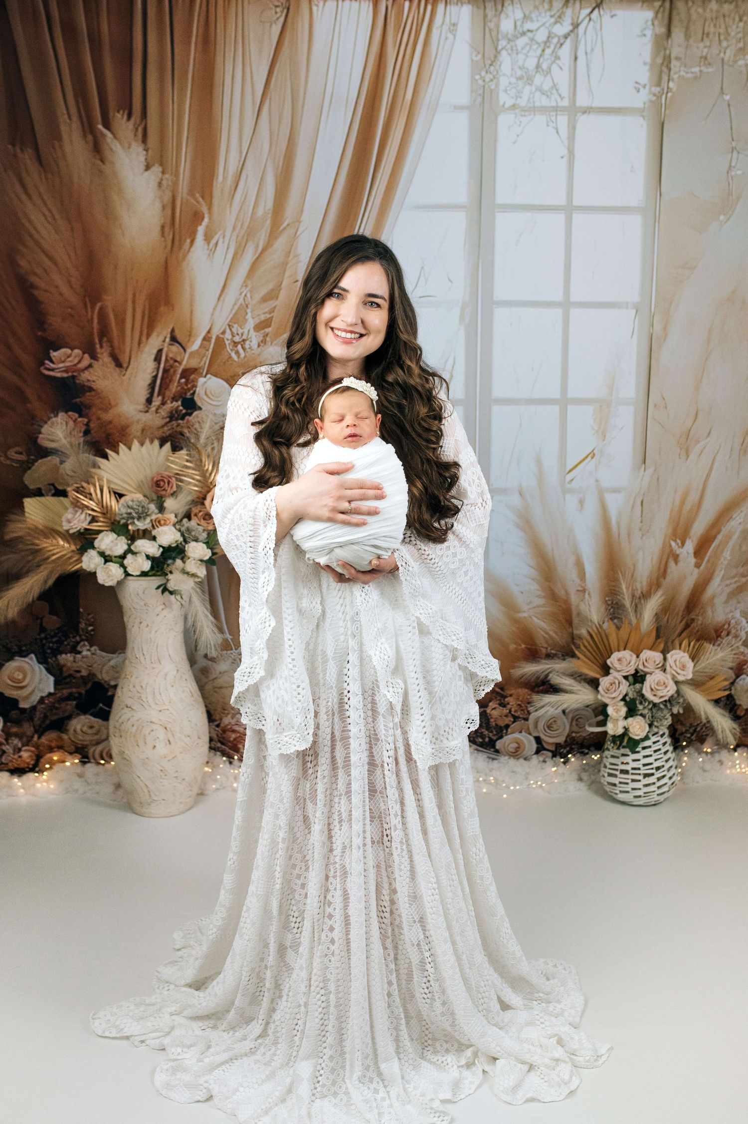 Woman in white lace gown holding swaddled newborn baby in boho floral studio setting.
