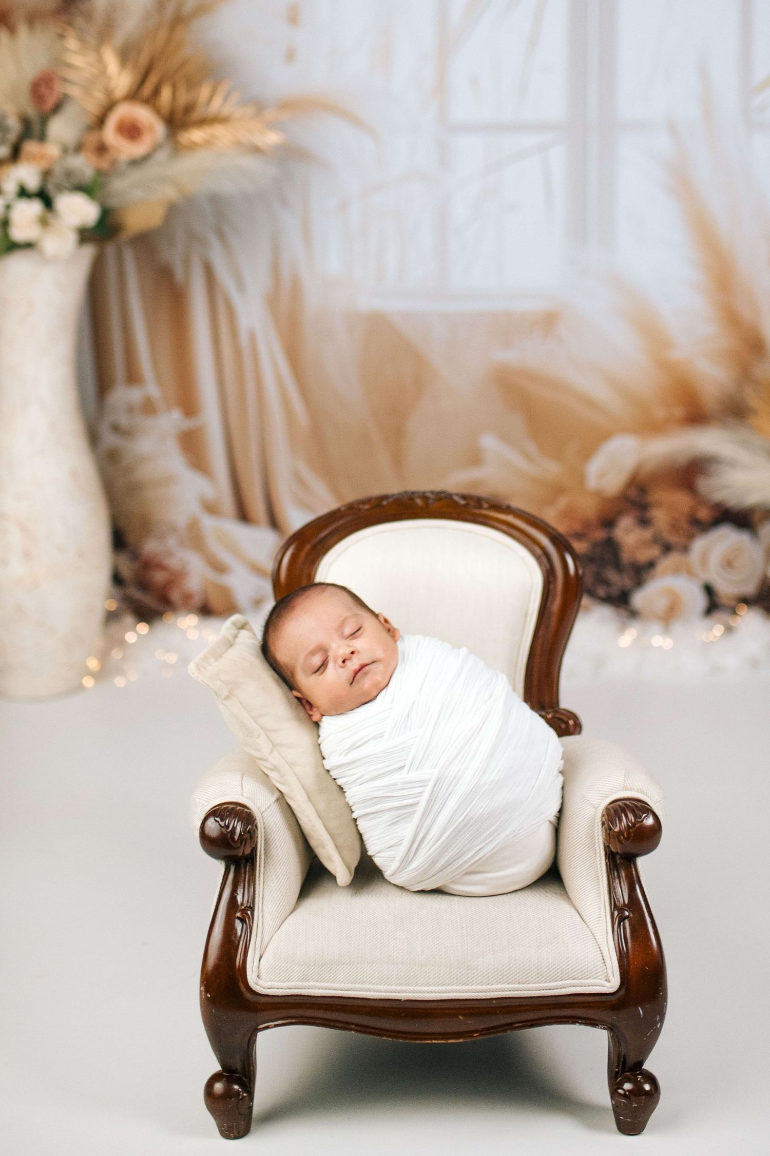Newborn baby swaddled in white wrap sleeping peacefully on a miniature cream armchair in a boho studio setting.
