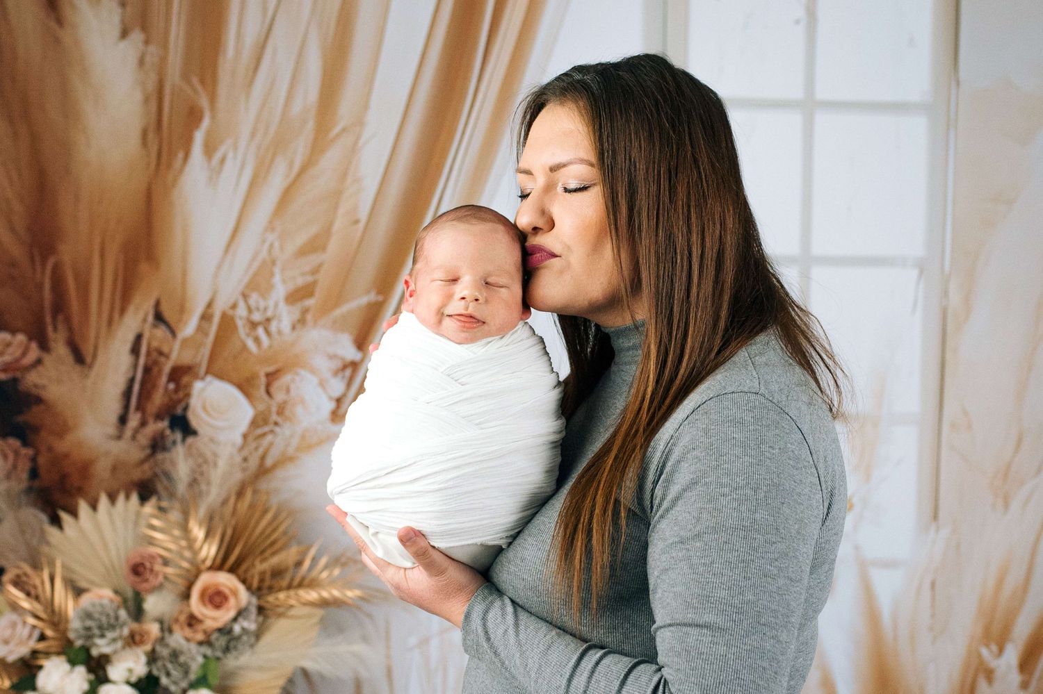 A mother tenderly kisses her swaddled newborn baby wrapped in white, surrounded by dried floral arrangements.