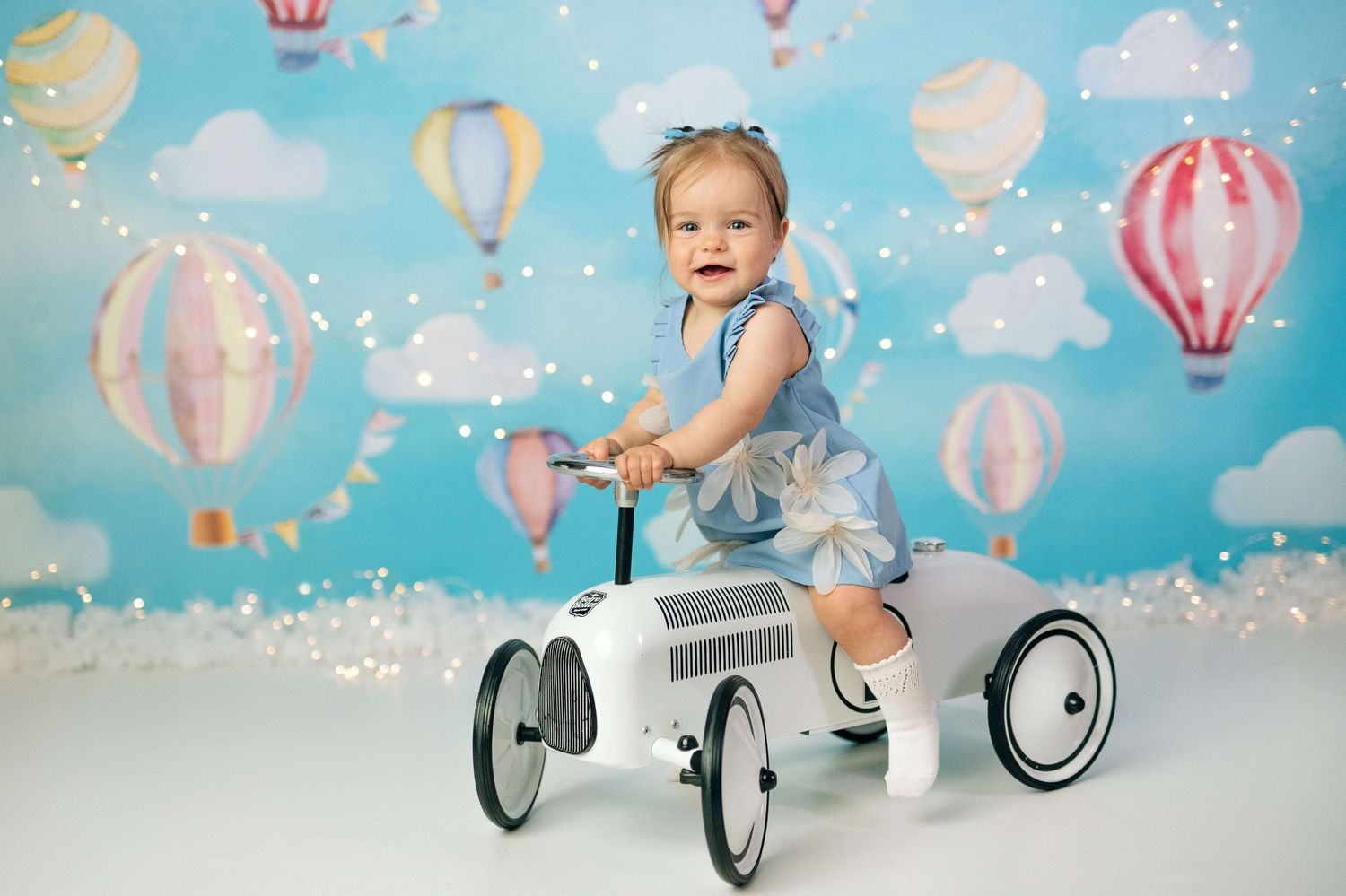 Toddler in blue dress riding a toy car in front of a colorful hot air balloon backdrop with twinkling lights.