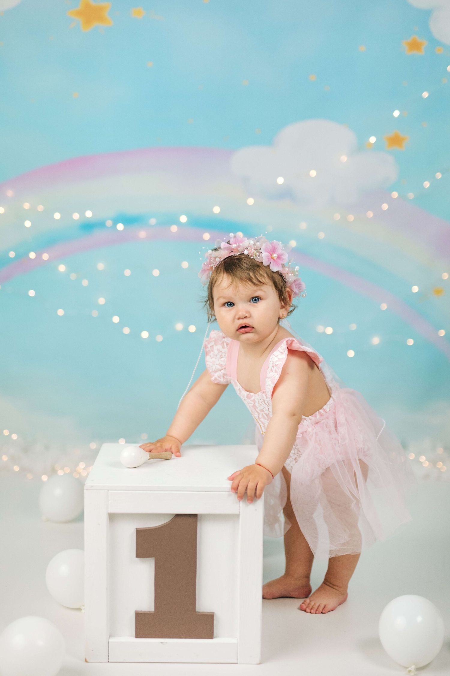 Baby girl in pink tutu and floral crown stands at a number one block, celebrating her first birthday with rainbow backdrop.