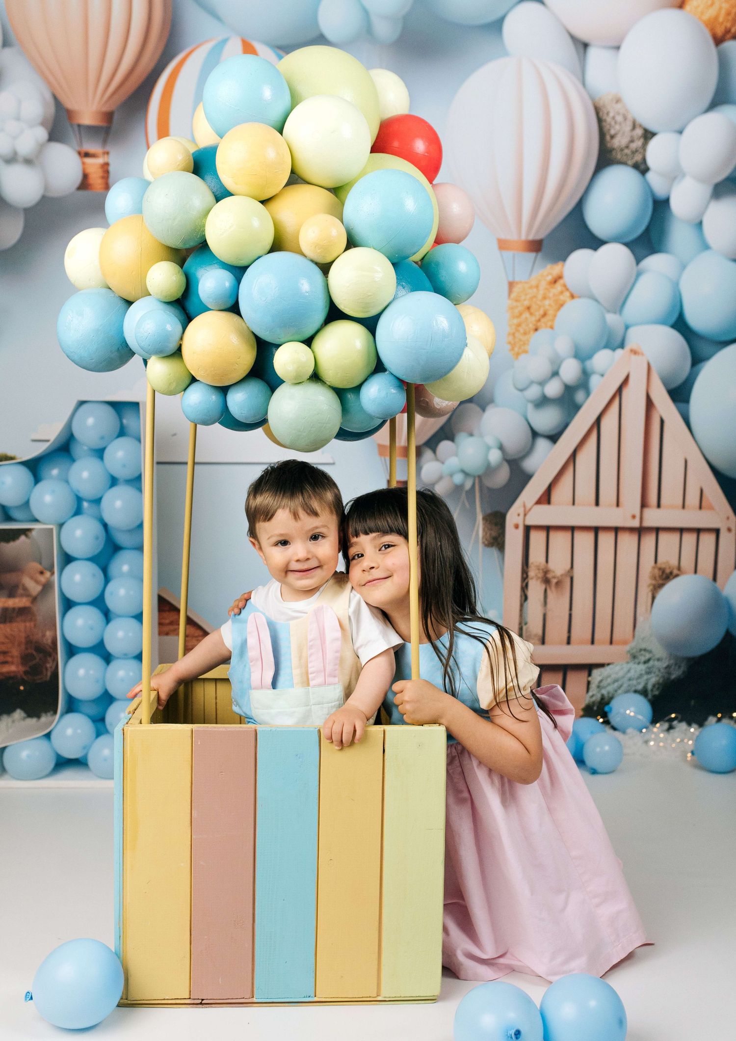 Two children pose in a colorful hot air balloon prop surrounded by pastel blue, yellow, and green balloons for a birthday photo.