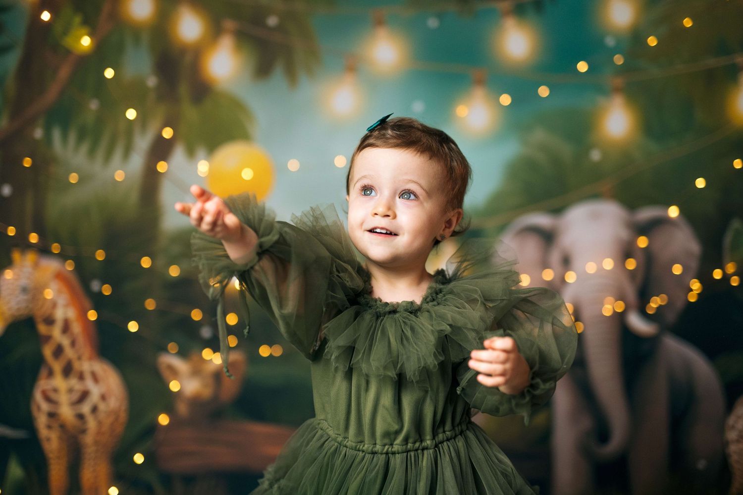 A toddler girl in a green tulle dress reaches for a glowing orb amid bokeh lights and jungle animal backdrop.