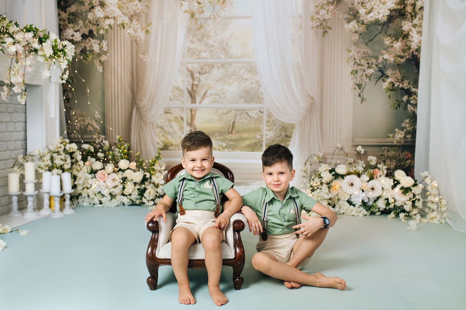 Two young boys in matching green shirts sit together on a vintage chair surrounded by white floral decorations.
