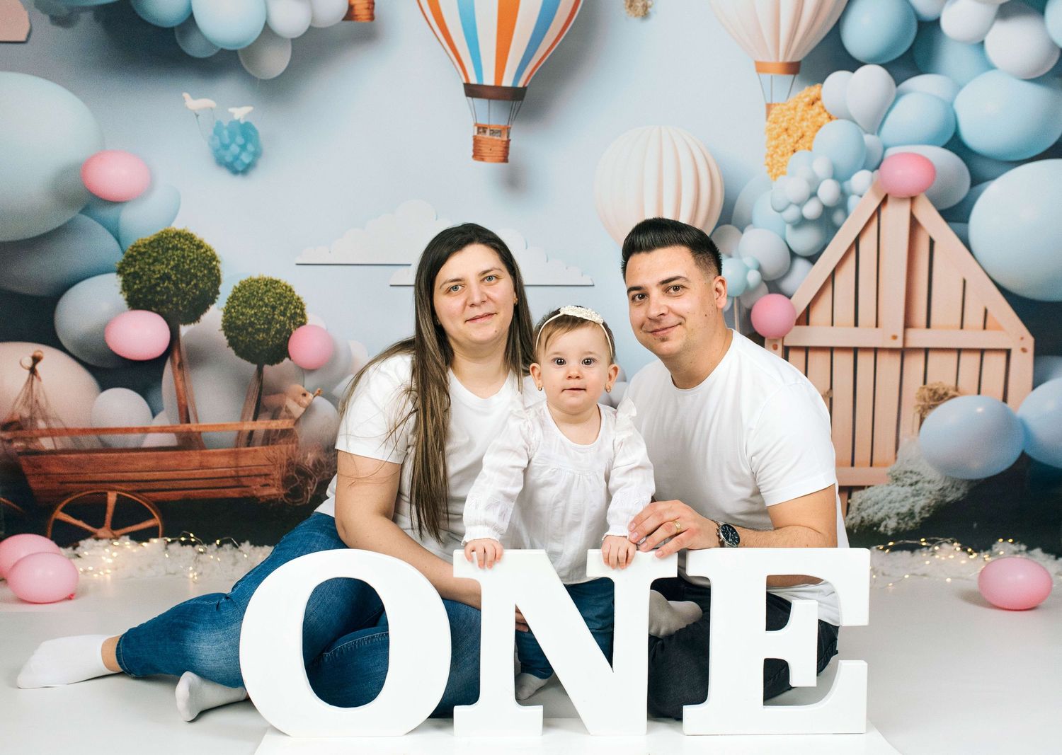 Family of three posing with ONE letters in front of colorful balloon and hot air balloon backdrop for first birthday.
