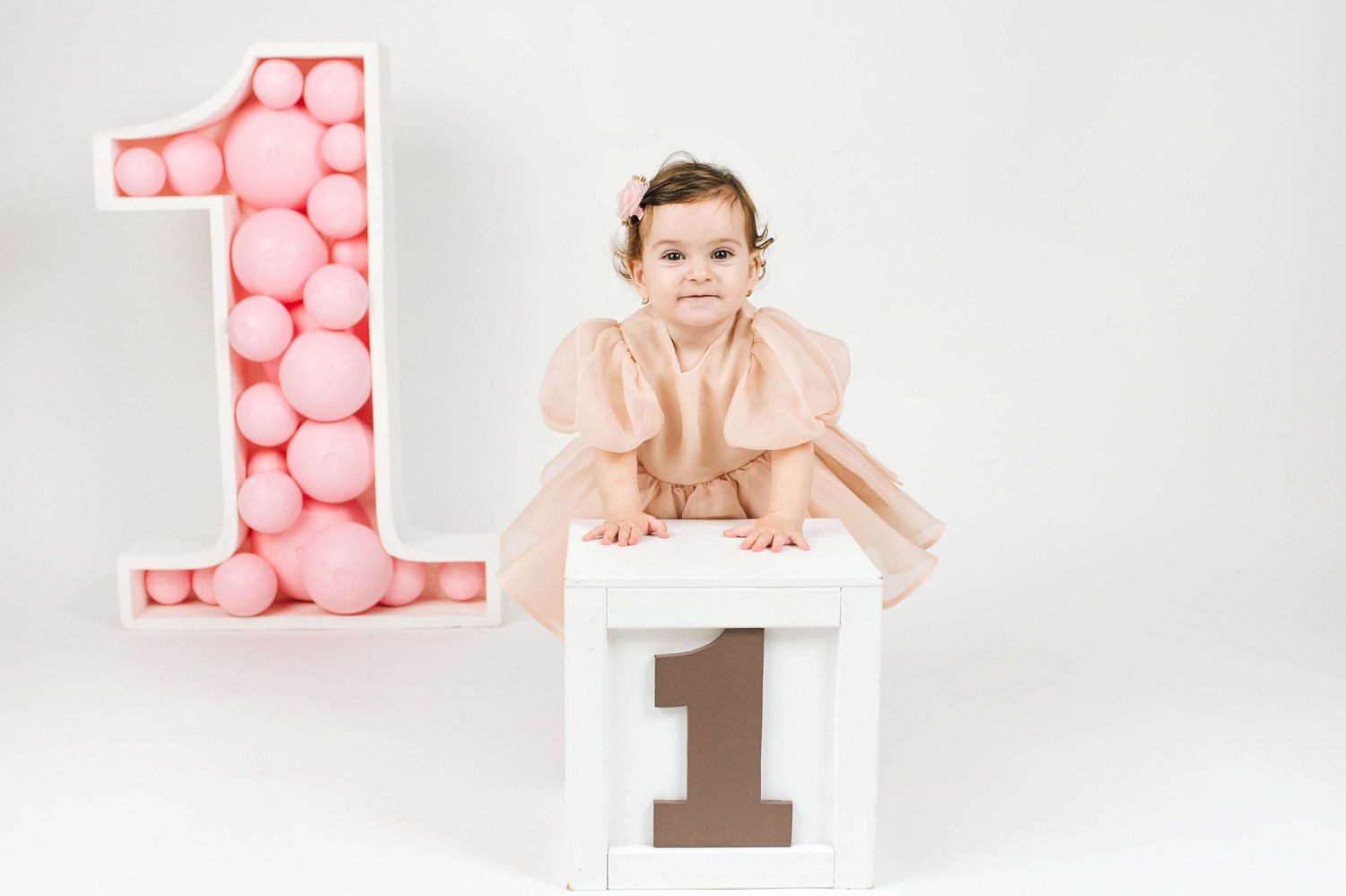Baby girl in pink dress poses with number one balloon decor for first birthday photo shoot.