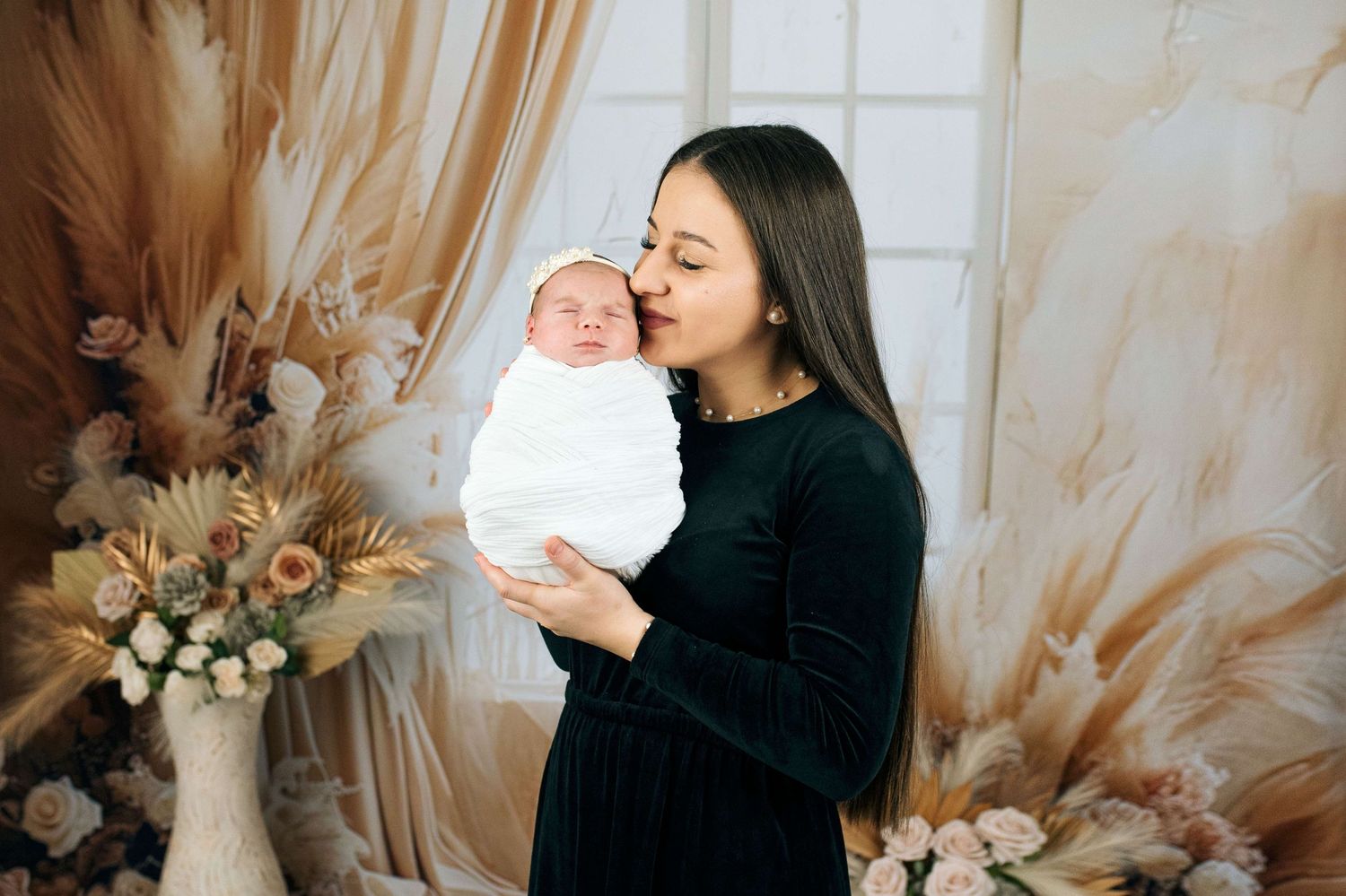 A woman in a black dress tenderly kisses a swaddled newborn baby amid floral decor.