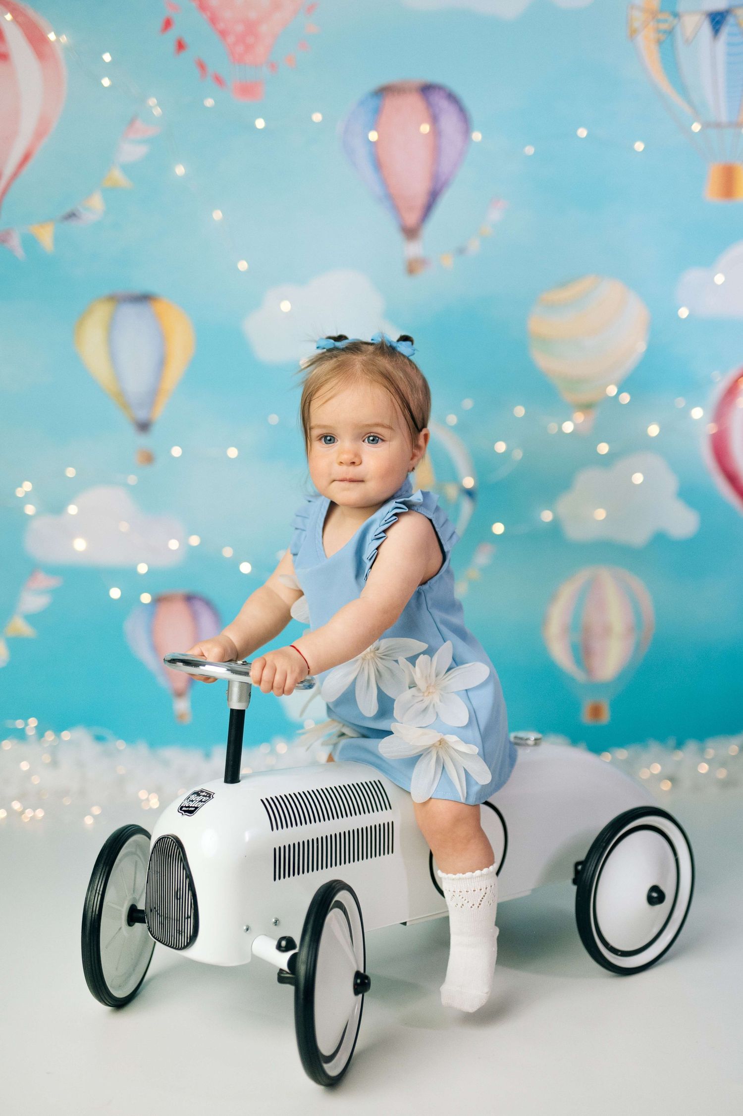 Toddler girl in blue dress riding white toy car against colorful hot air balloon backdrop.