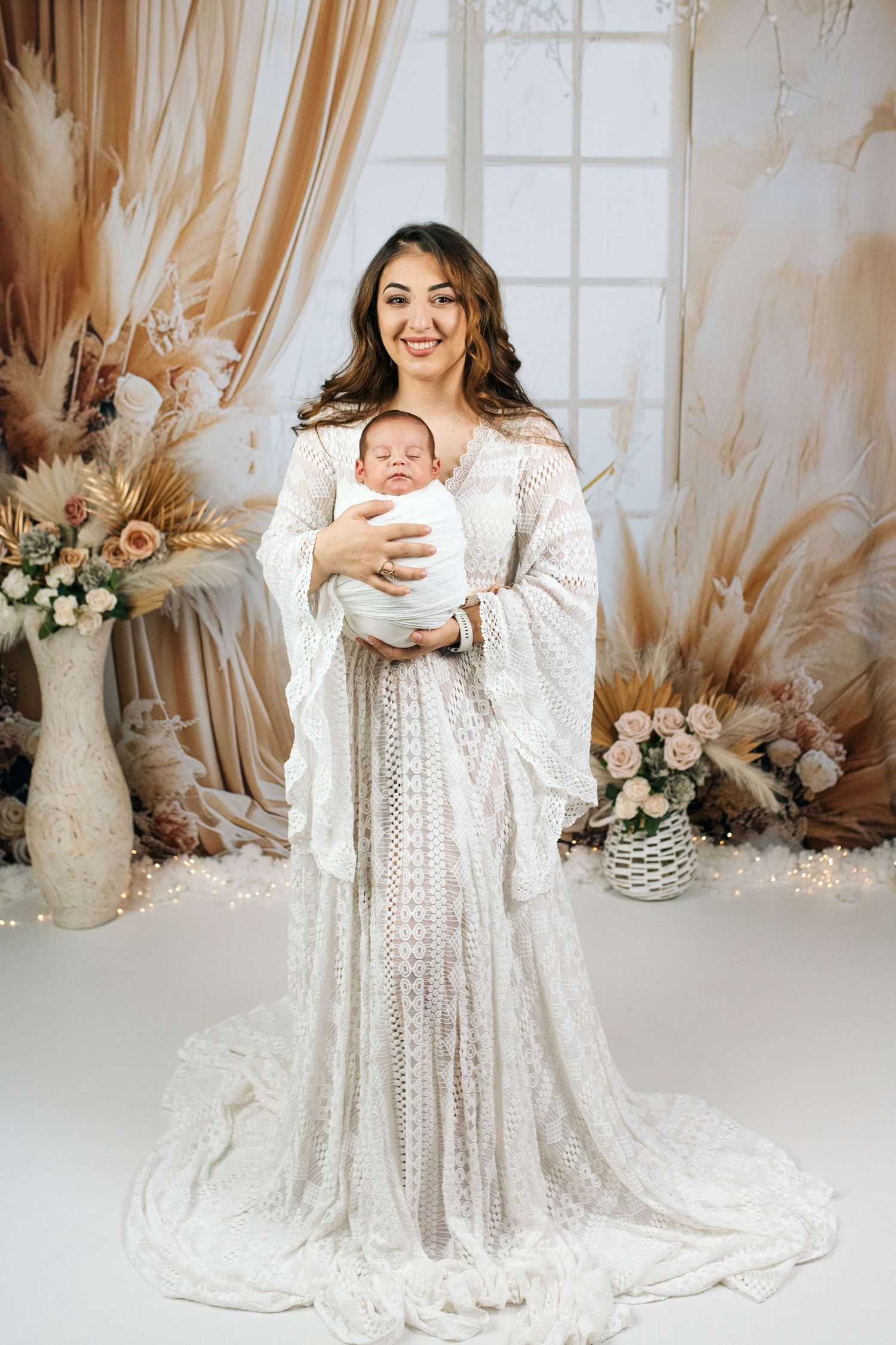 A smiling woman in a white lace boho gown holds a swaddled newborn in a floral studio setting.