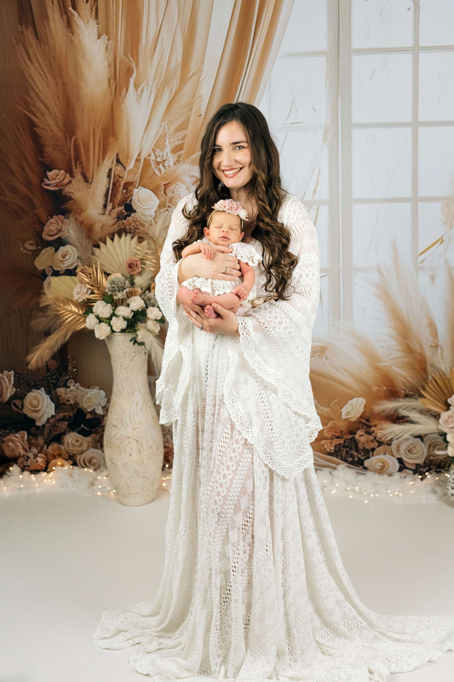 Smiling woman in white lace boho gown holding newborn baby in floral studio setting.
