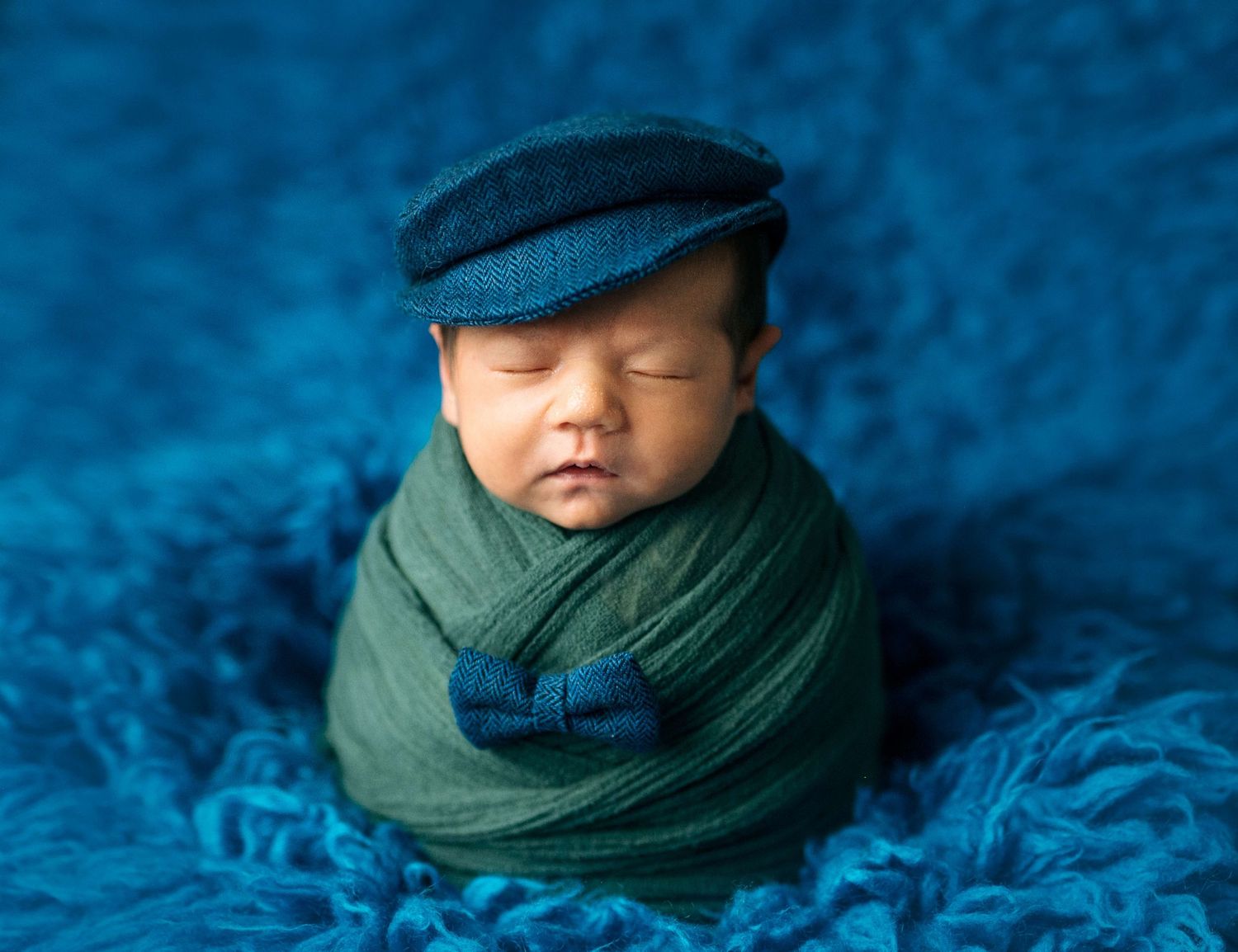 Sleeping newborn baby wrapped in green swaddle wearing a navy blue flat cap on a blue fluffy blanket.