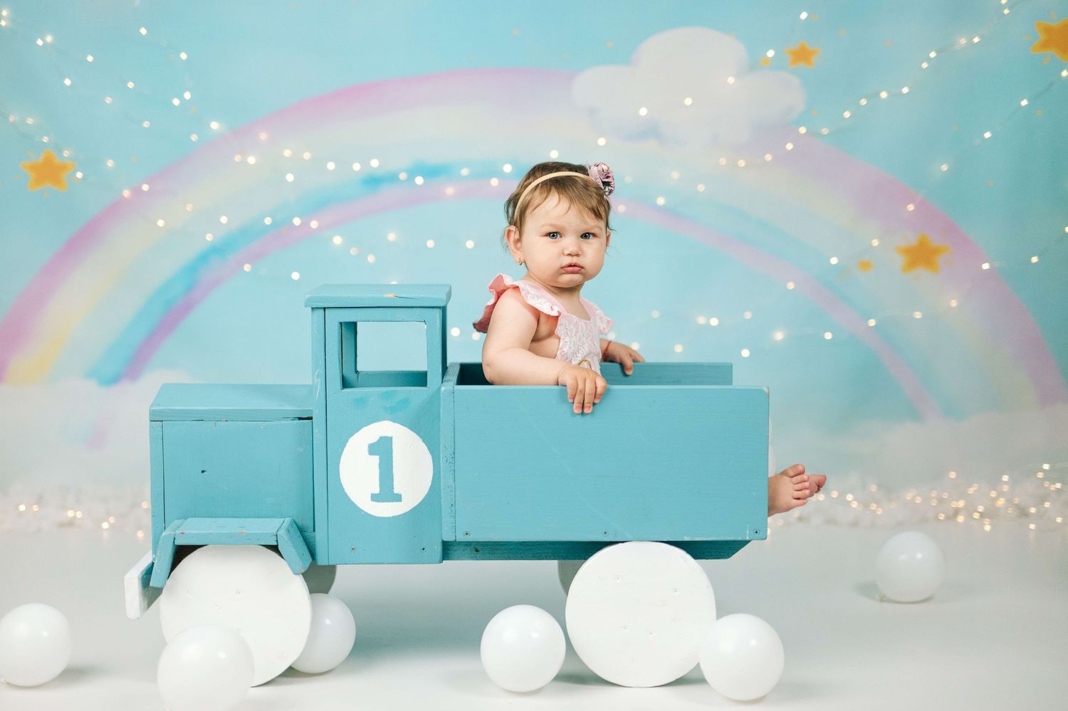 Baby girl sits in a blue toy truck with a rainbow backdrop for her first birthday cake smash photo session.