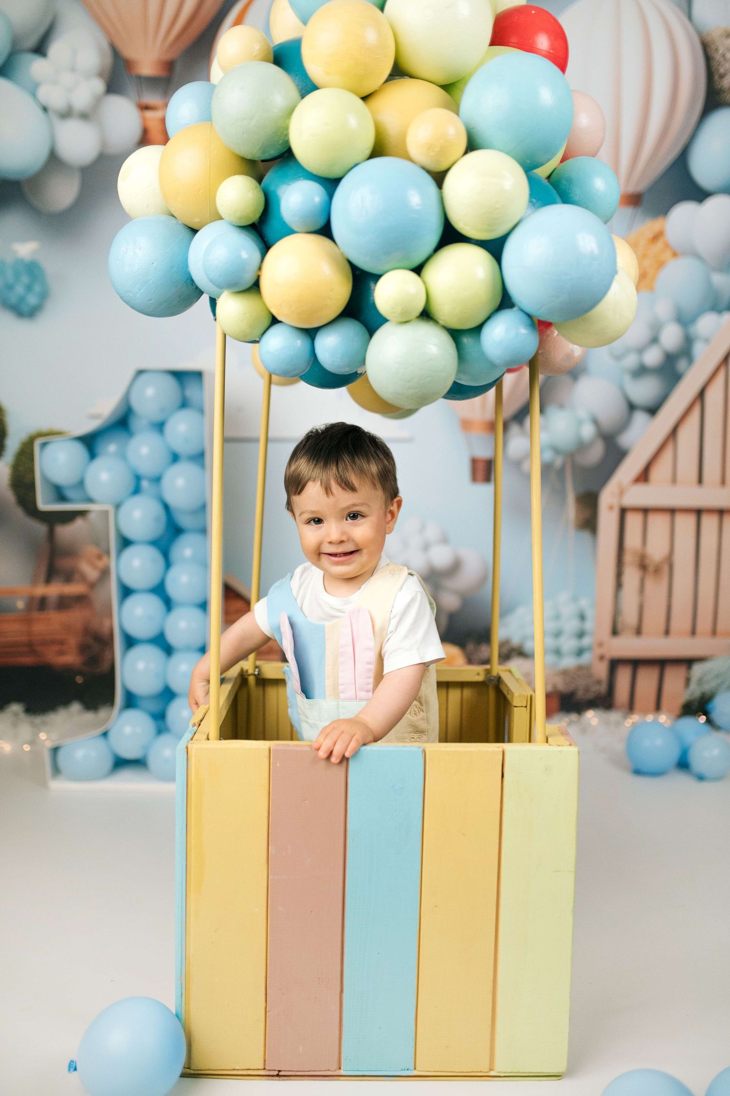 Smiling toddler standing in a colorful hot air balloon prop surrounded by blue and yellow balloons at a first birthday party.