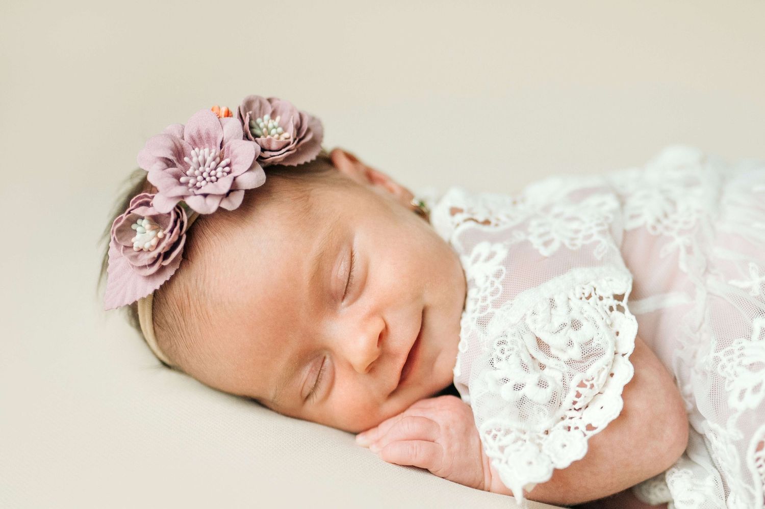 Sleeping newborn baby girl wearing a pink floral headband and white lace wrap in a professional portrait photo.