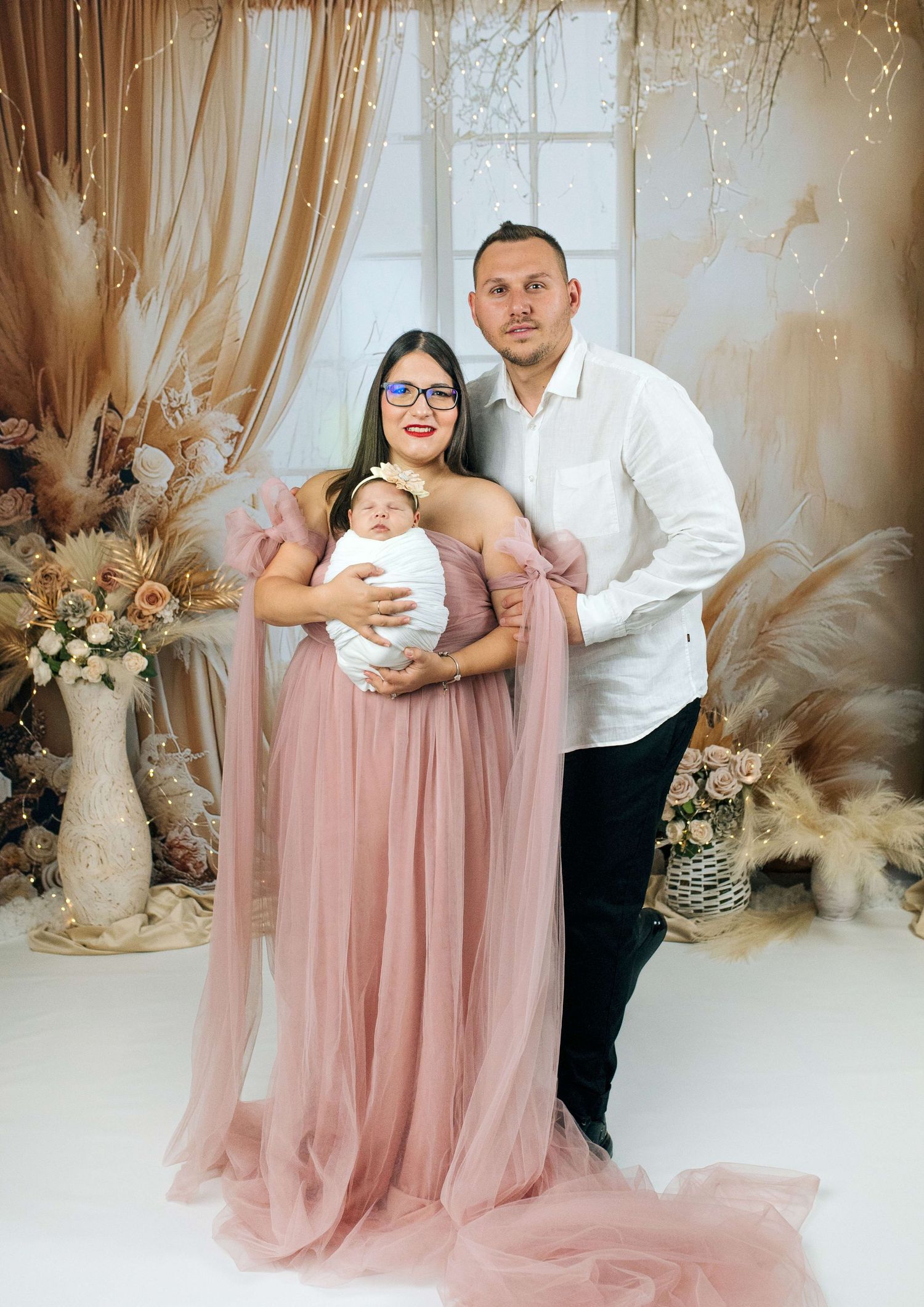 A couple in elegant attire holds their newborn baby in a beautifully decorated studio with floral arrangements.