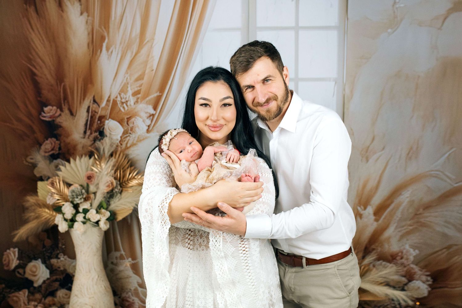A happy couple in elegant attire holds their newborn twins in a beautifully decorated studio with floral arrangements.