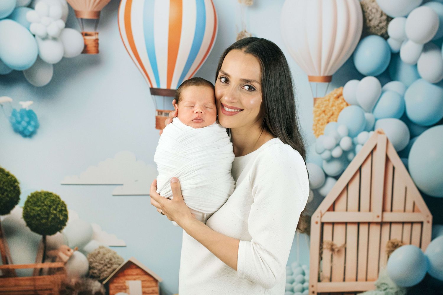 A young mother with dark hair holds her swaddled newborn baby against a blue balloon-themed backdrop.