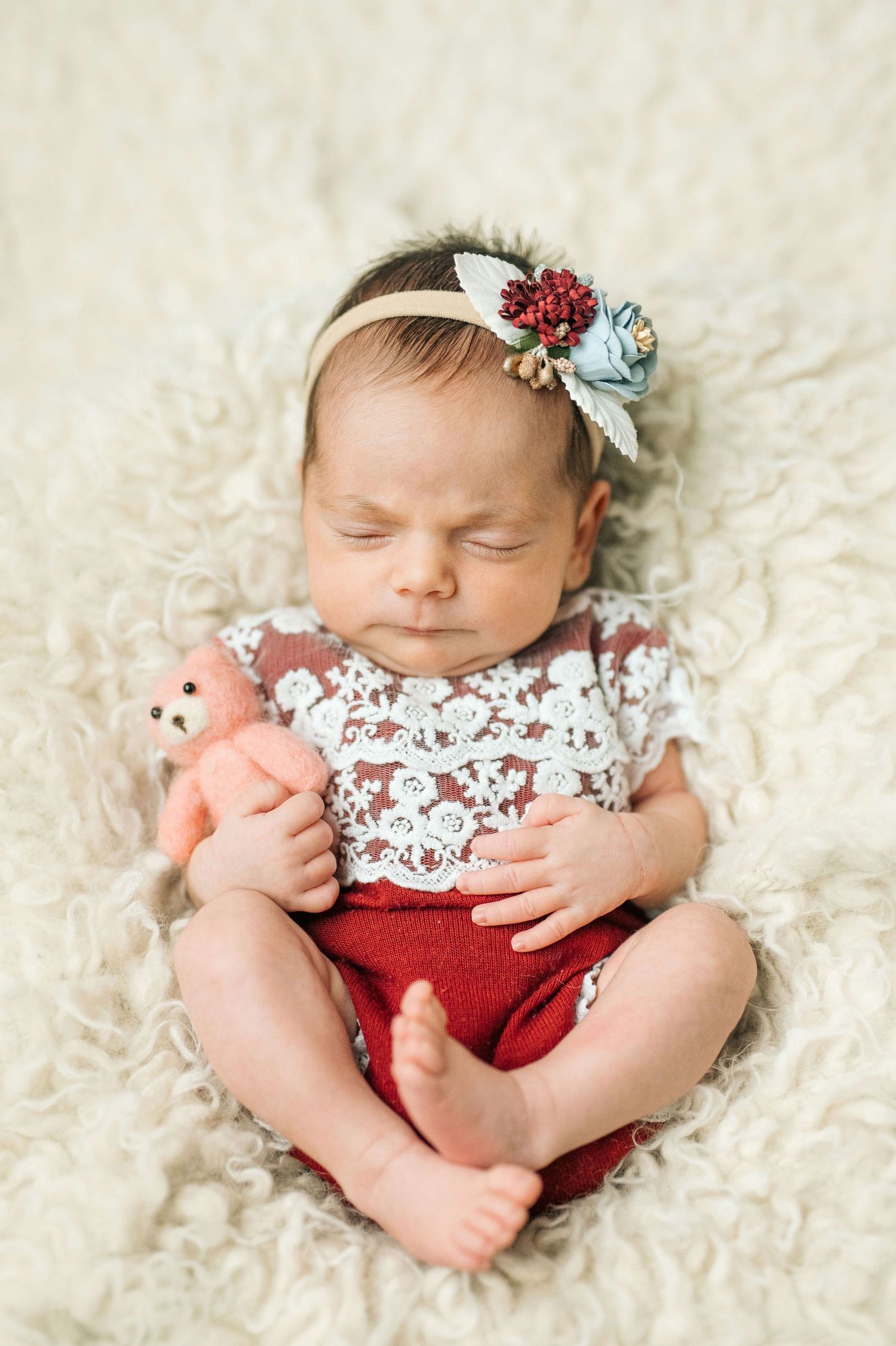 Sleeping newborn baby girl in lace outfit and floral headband holding a pink teddy bear on fluffy white blanket.