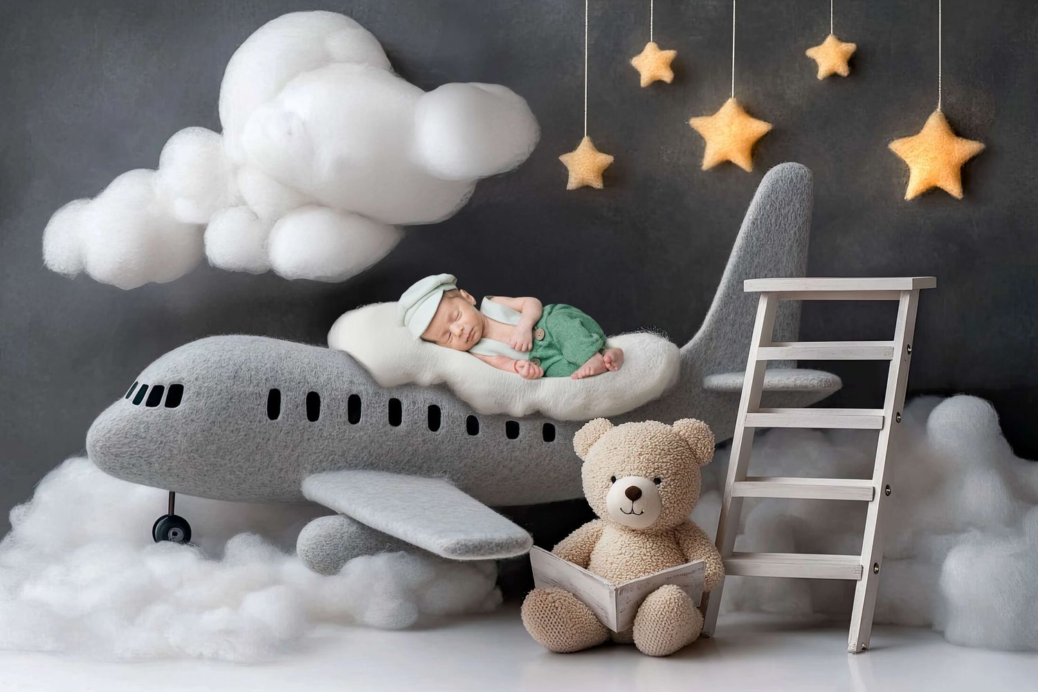 Newborn baby sleeping on a gray plush airplane with a teddy bear, clouds, and hanging stars on a dark backdrop.