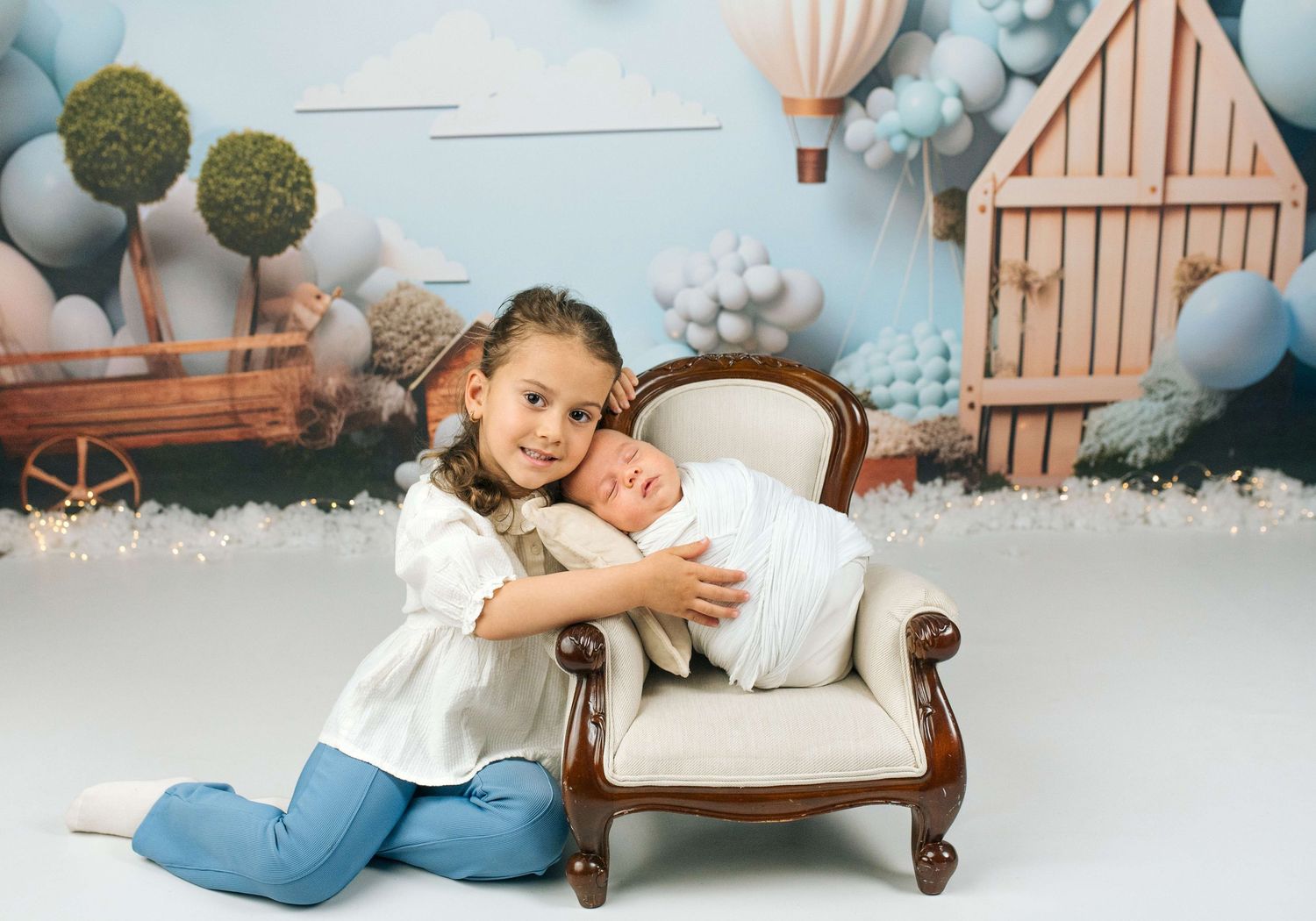 Young girl in white top and jeans hugs sleeping newborn on mini armchair with balloon backdrop.