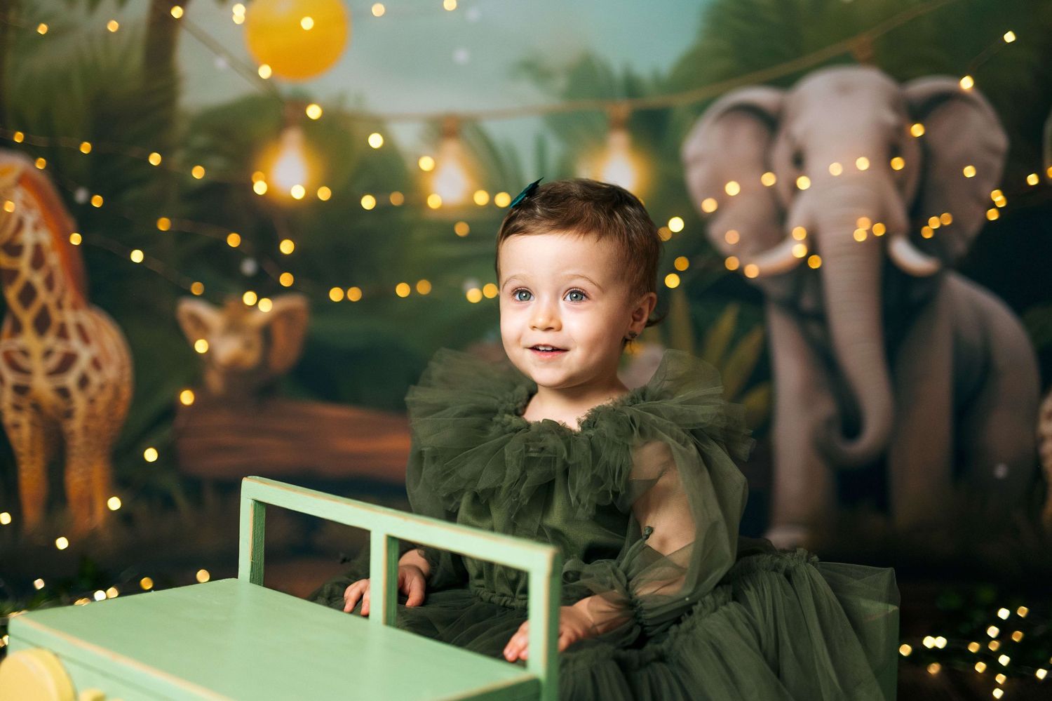 Toddler in green tulle dress poses at mini table with safari animal backdrop and bokeh lights.