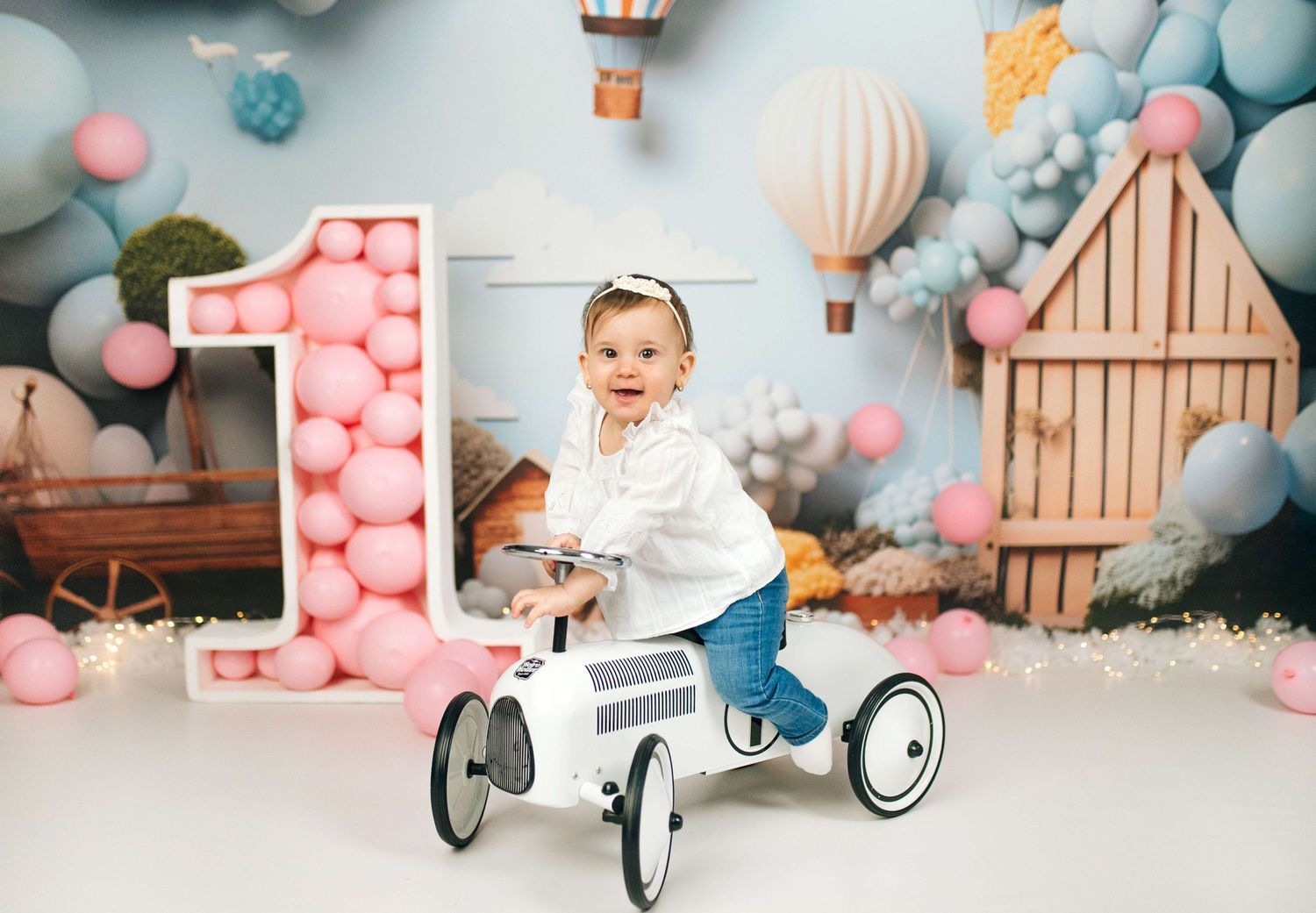 Baby girl on toy car at 1st birthday cake smash with pink balloons and hot air balloon backdrop.