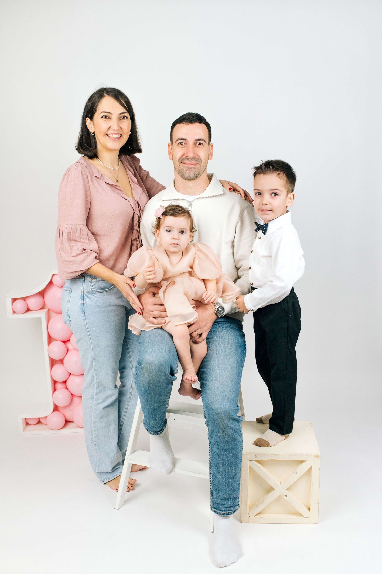 Family of four posing for a first birthday celebration photo with pink balloon number one decoration.